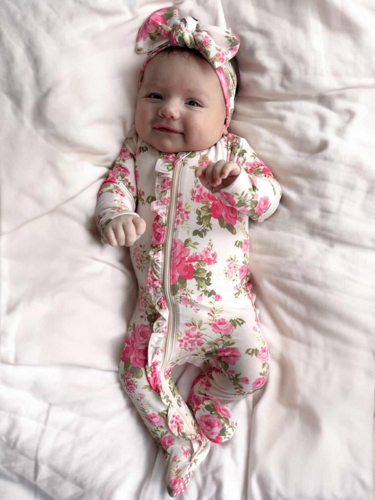 Smiling baby in floral onesie and matching headband, lying on soft white bedding.