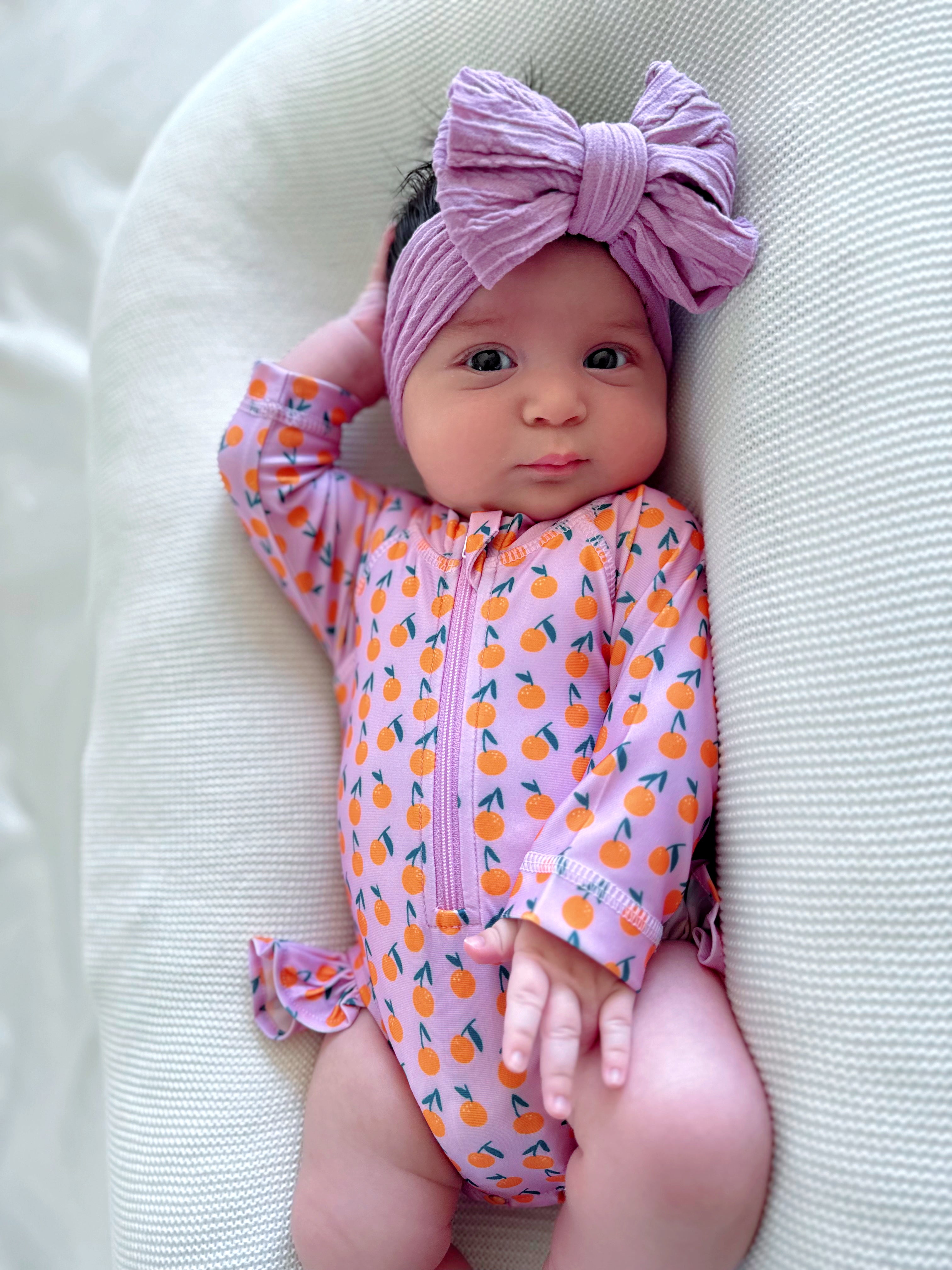 Baby wearing a colorful orange-patterned romper and a large lavender headband, resting against a soft background.