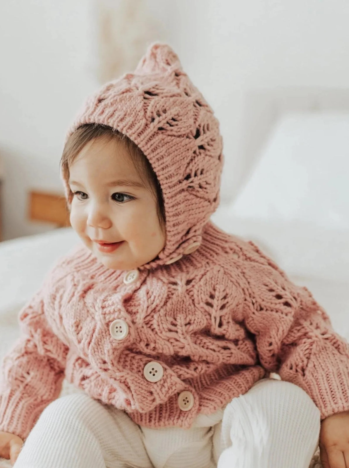 Smiling child in a pink knitted sweater and hat, seated on a bed in a cozy room.