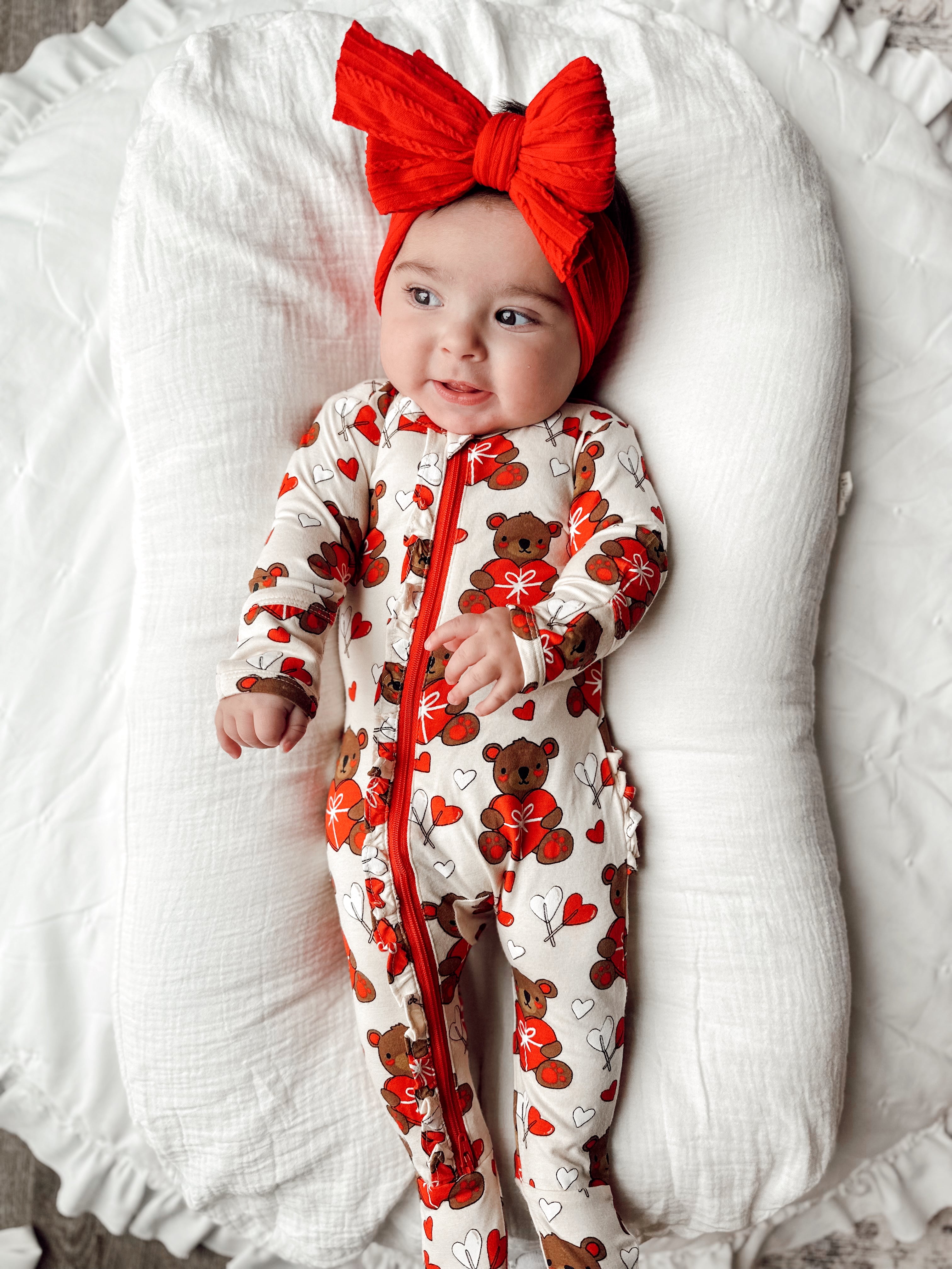 Smiling baby in a bear-themed onesie and red bow headband, lying on a plush white blanket.