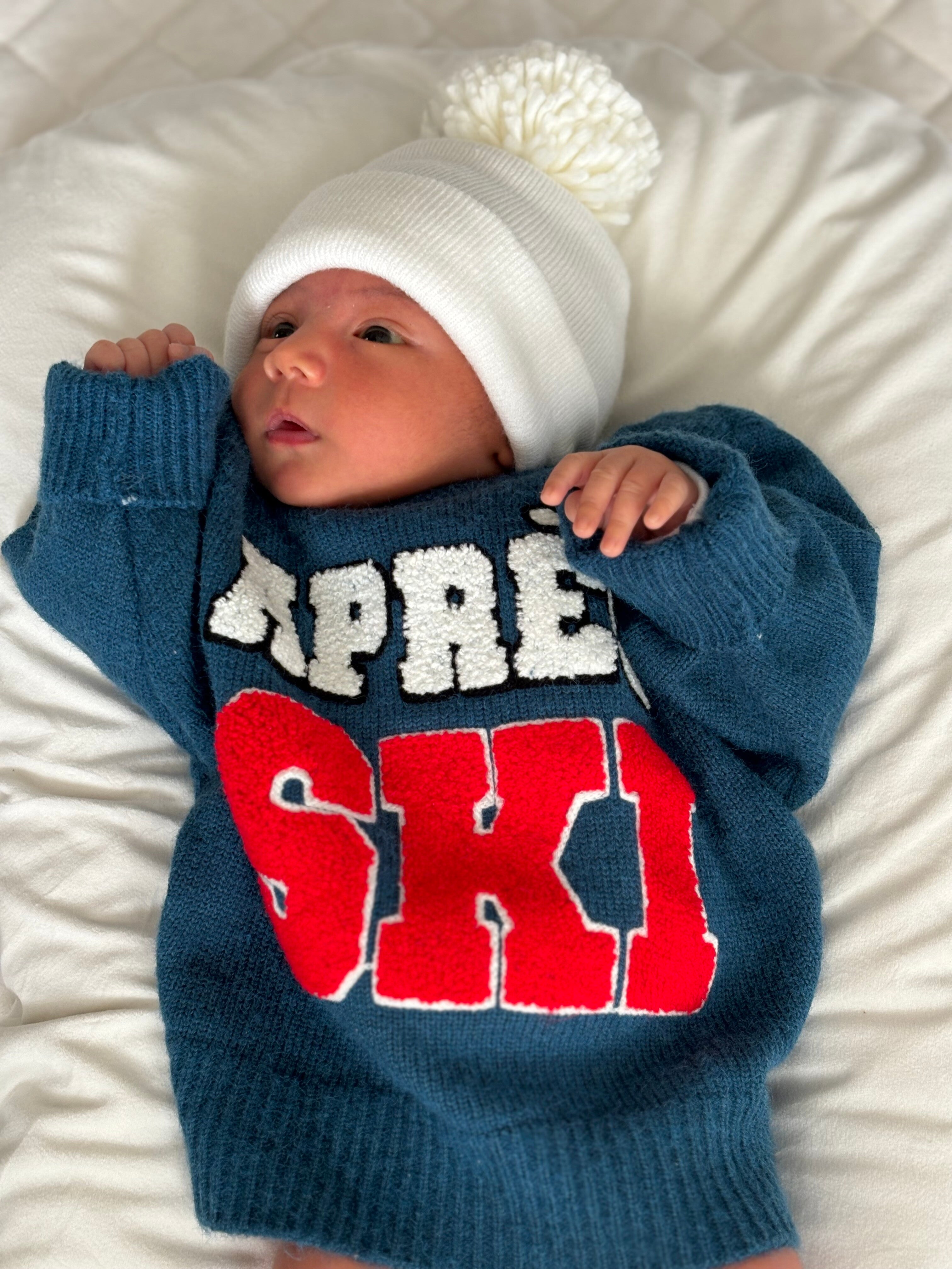 Baby wearing a blue sweater with "Supreme Ski" in white and red, and a white pom-pom beanie, lying on a white blanket.