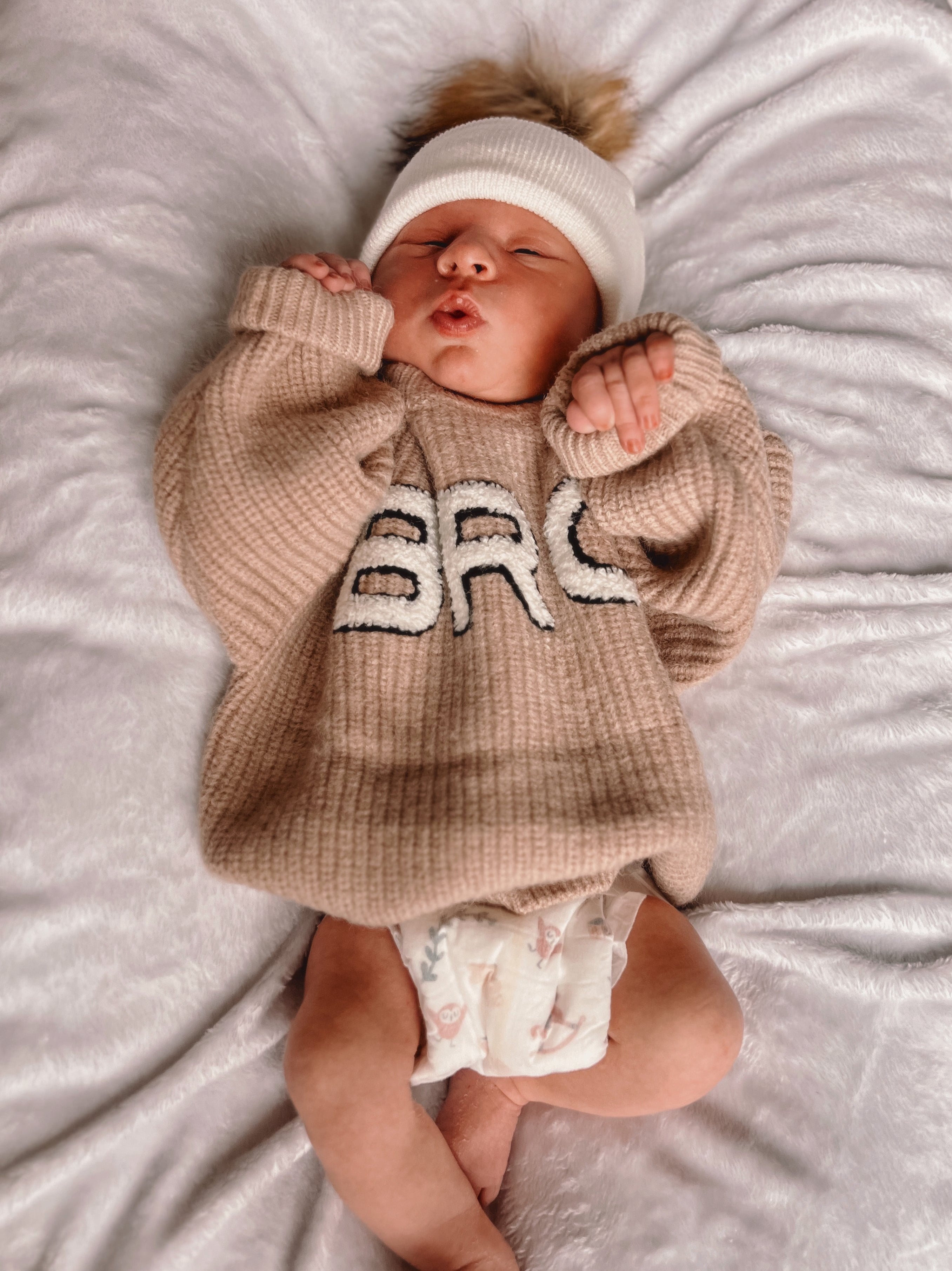 Newborn baby in a cozy beige sweater with "BRC" lettering, lying on a fluffy blanket, wearing a white hat.