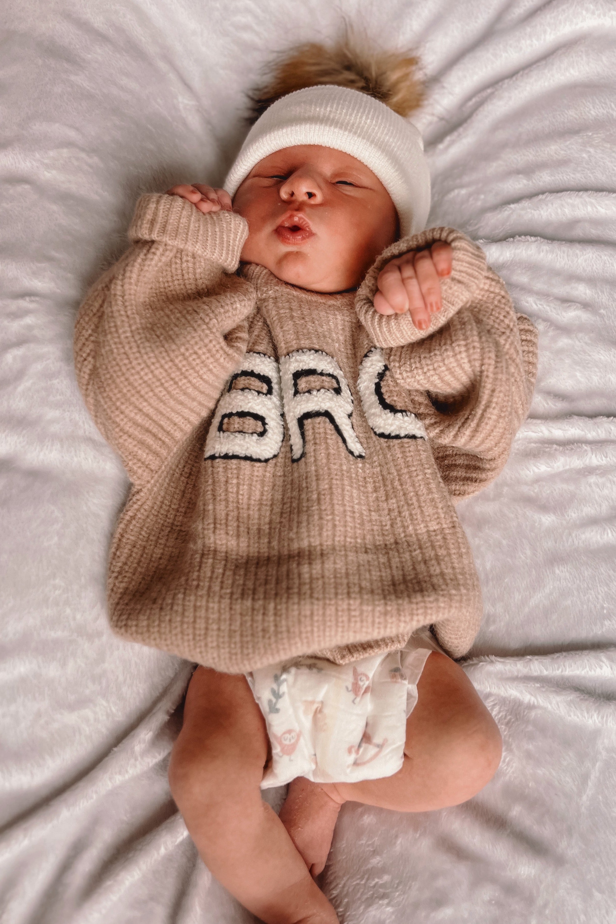 Newborn baby in a cozy beige sweater with "BRC" lettering, lying on a fluffy blanket, wearing a white hat.