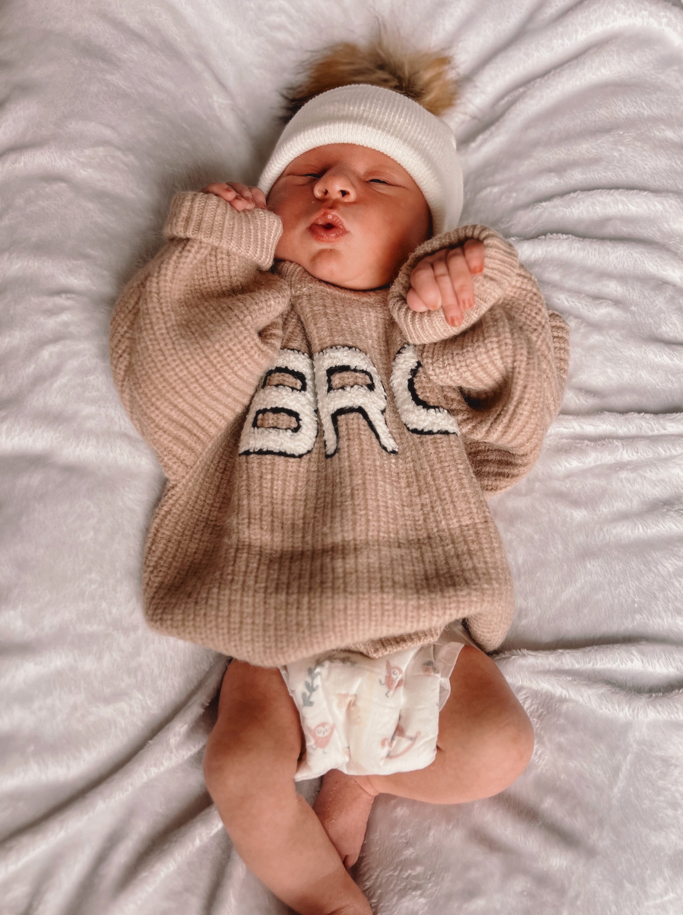 Newborn baby in a cozy beige sweater with "BRC" lettering, lying on a fluffy blanket, wearing a white hat.