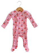 Pink baby onesie with cherry prints, featuring long sleeves and a zip closure, hung on a wooden hanger.