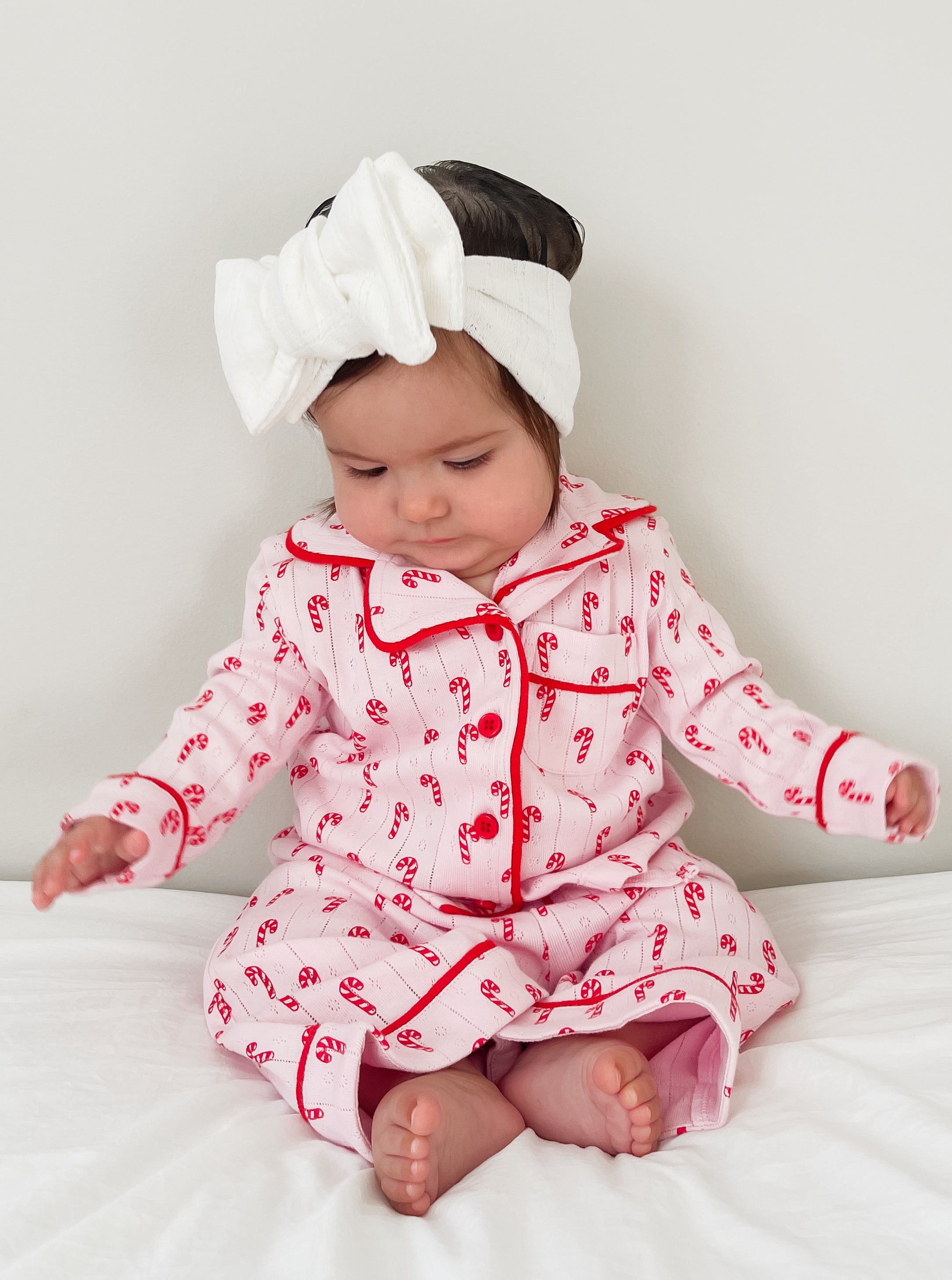 Baby in holiday-themed pajamas with candy cane print, wearing a white headband, sitting on a bed.