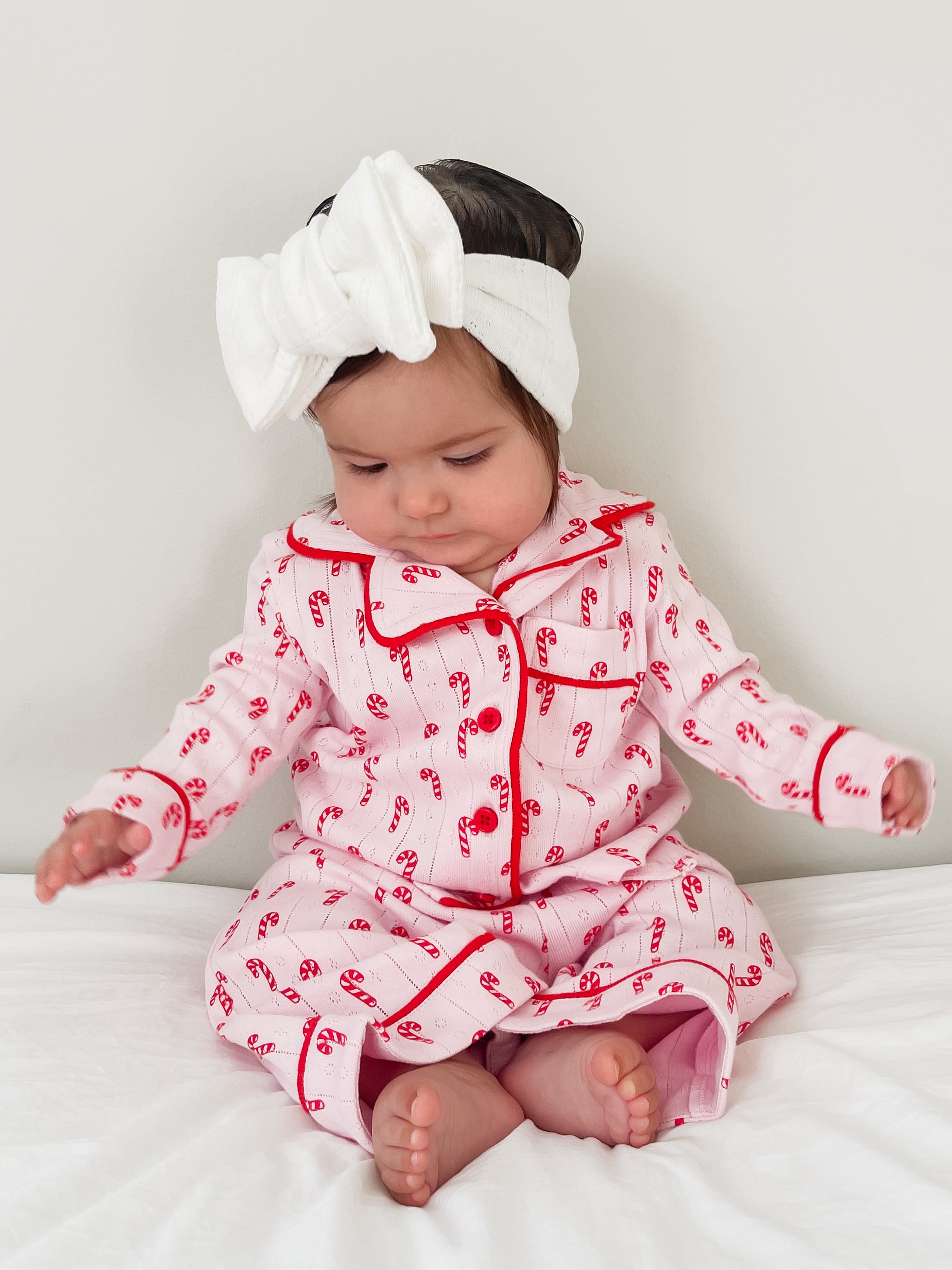 Baby in holiday-themed pajamas with candy cane print, wearing a white headband, sitting on a bed.