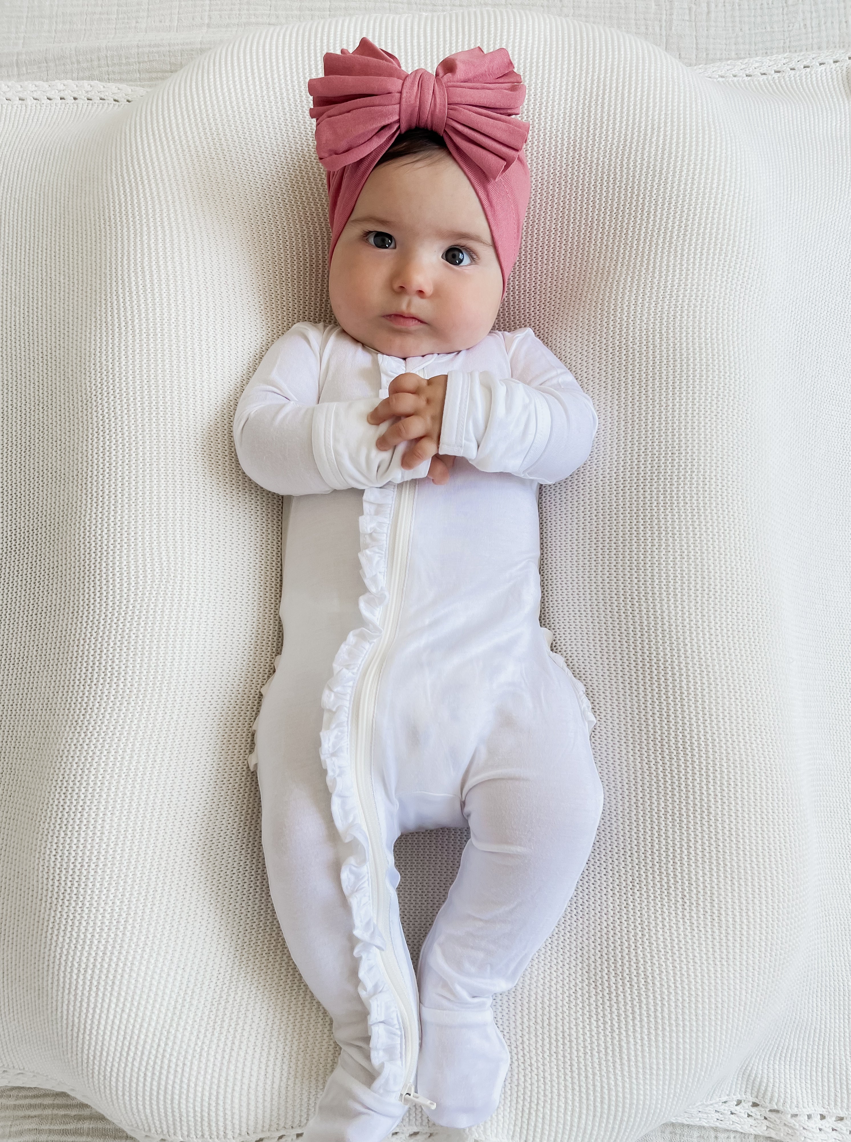 Baby in white onesie with ruffles, wearing a pink bow headband, resting on a textured white surface.