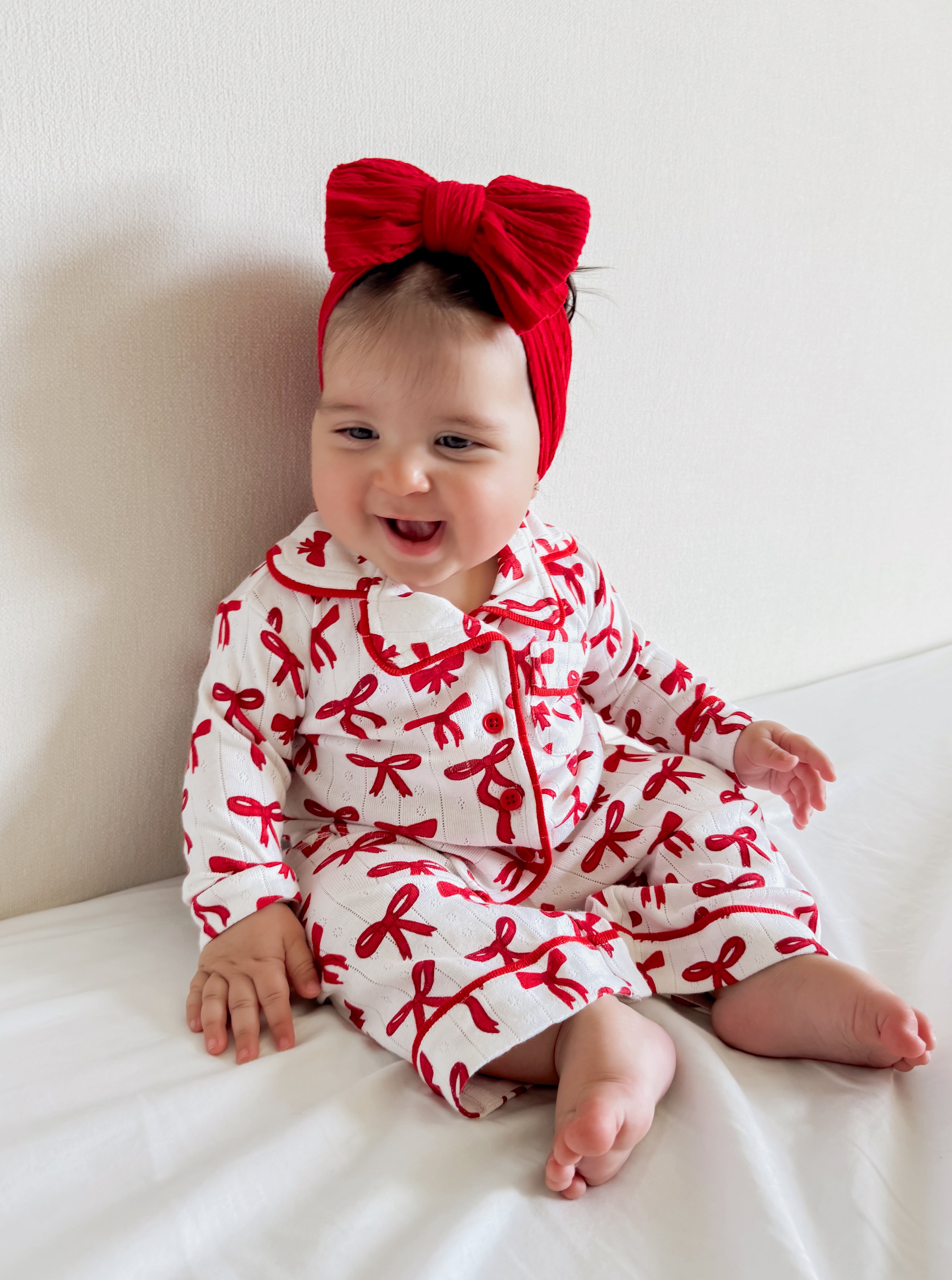 Smiling baby in a red bow headband and festive pajamas sitting on a bed against a light background.