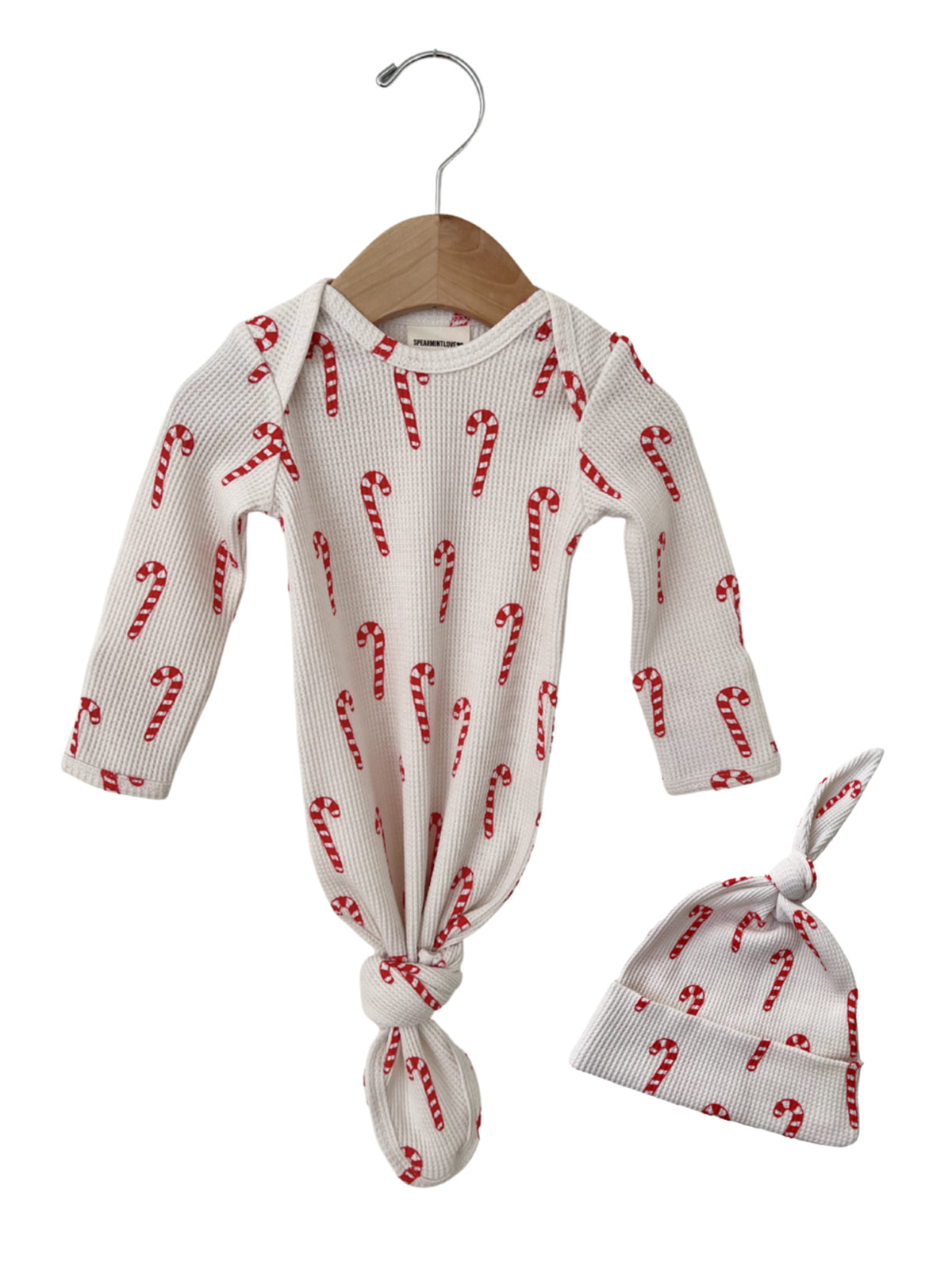 White baby outfit with red candy cane print, including a knotted hat and long-sleeve onesie, hanging on a wooden hanger.