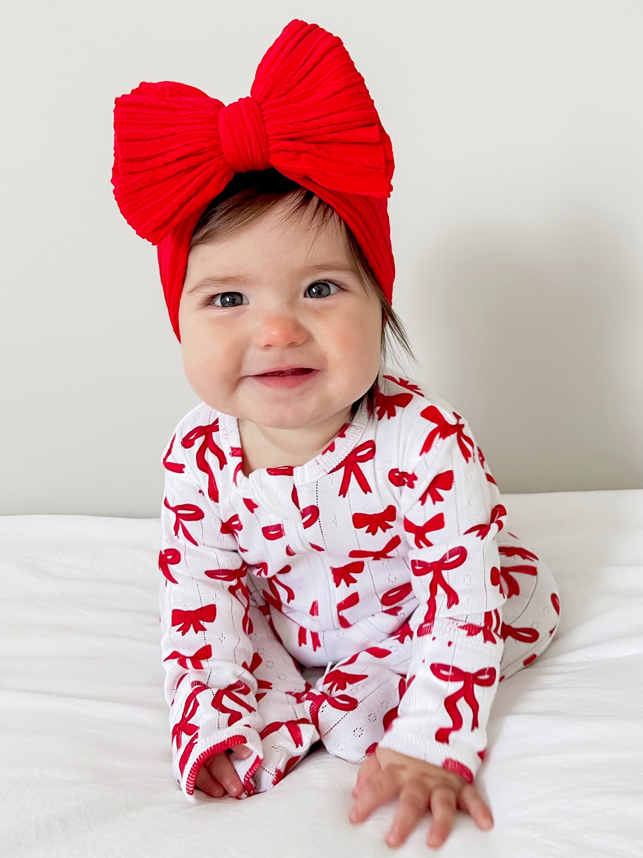 Smiling baby in a red bow headband and matching outfit with a white background.