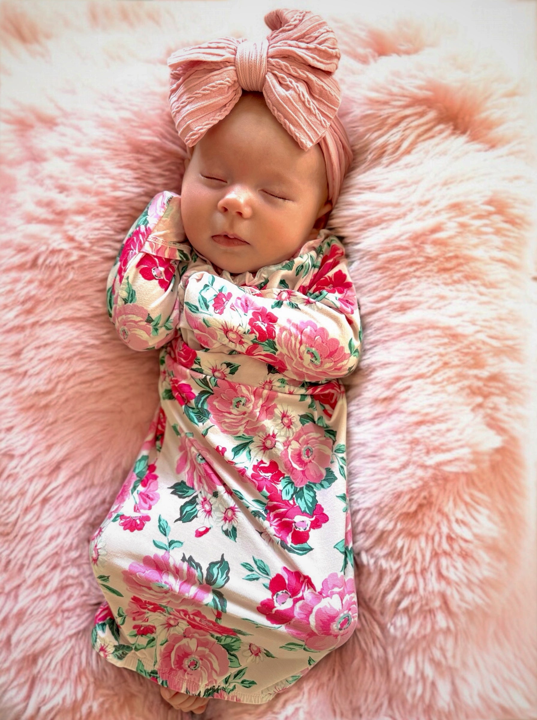 Sleeping baby girl in floral outfit, wearing a pink headband, resting on a soft pink fur blanket.
