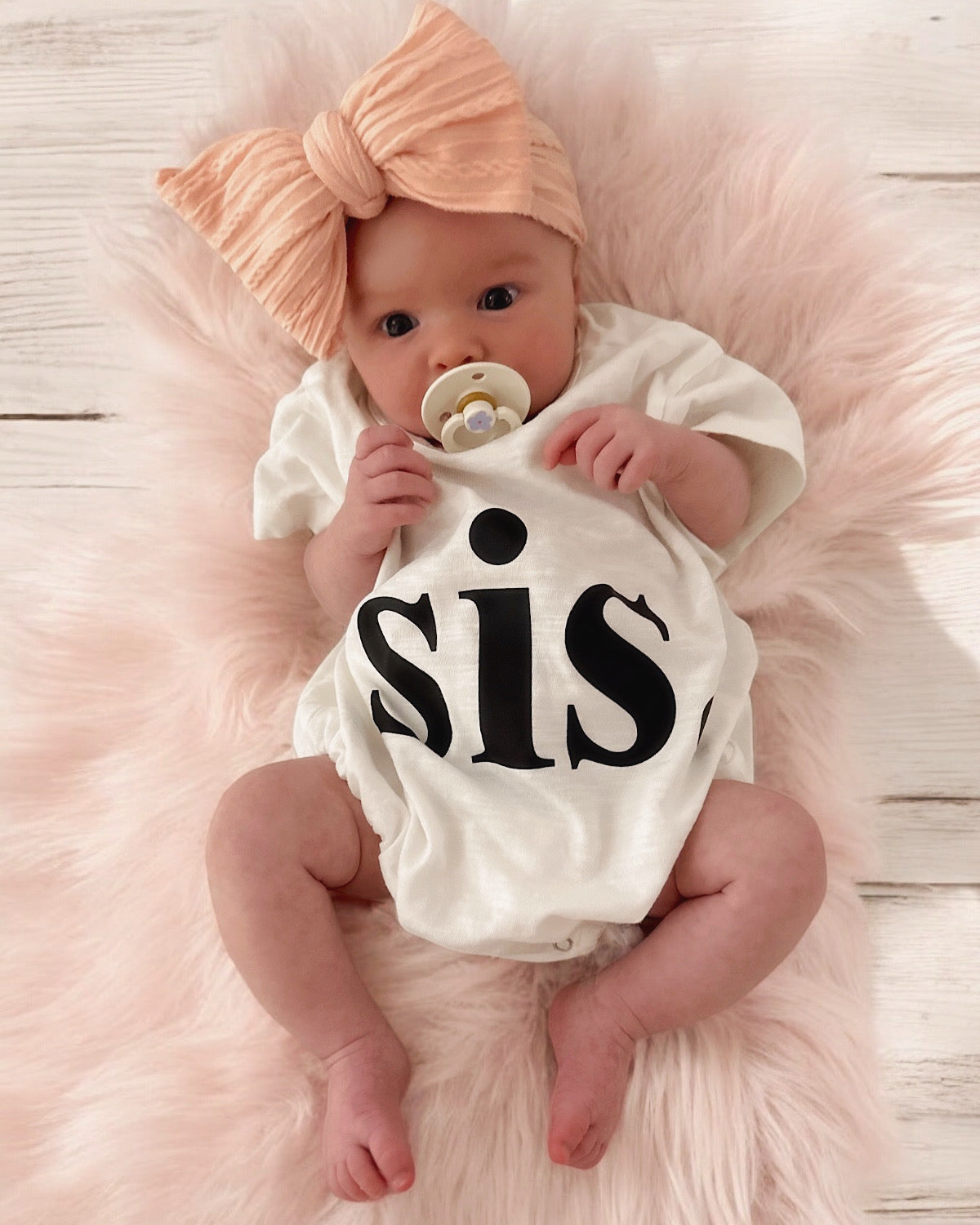 Baby girl in a "sis" onesie, wearing a large peach bow, sitting on a soft pink rug.