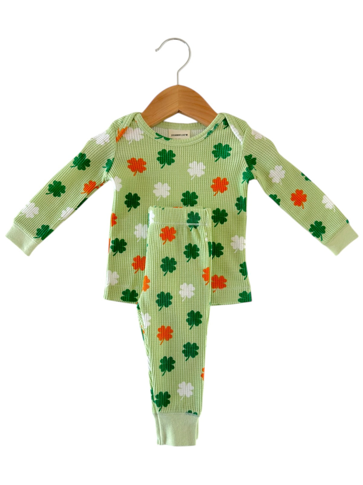 Green children's pajamas with shamrock patterns in green, white, and orange hanging on a wooden hanger.