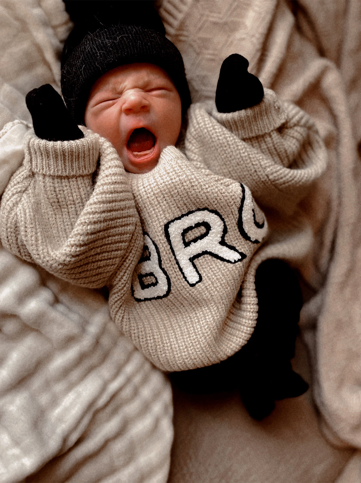 Yawning baby in a cozy sweater, wearing a black beanie and mittens, against a soft, textured background.