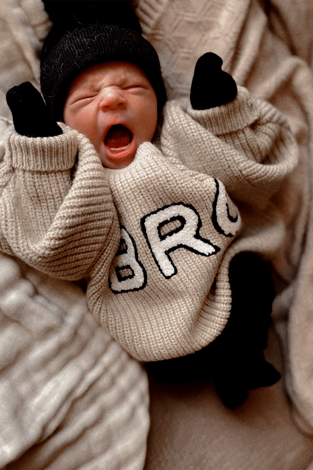 Yawning baby in a cozy sweater, wearing a black beanie and mittens, against a soft, textured background.