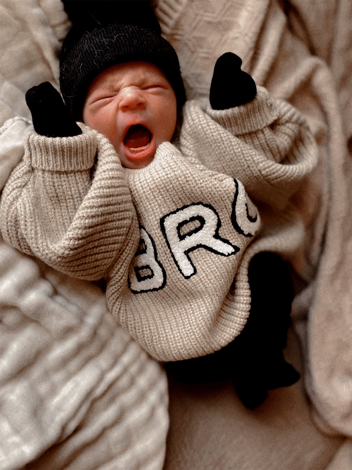 Yawning baby in a cozy sweater, wearing a black beanie and mittens, against a soft, textured background.