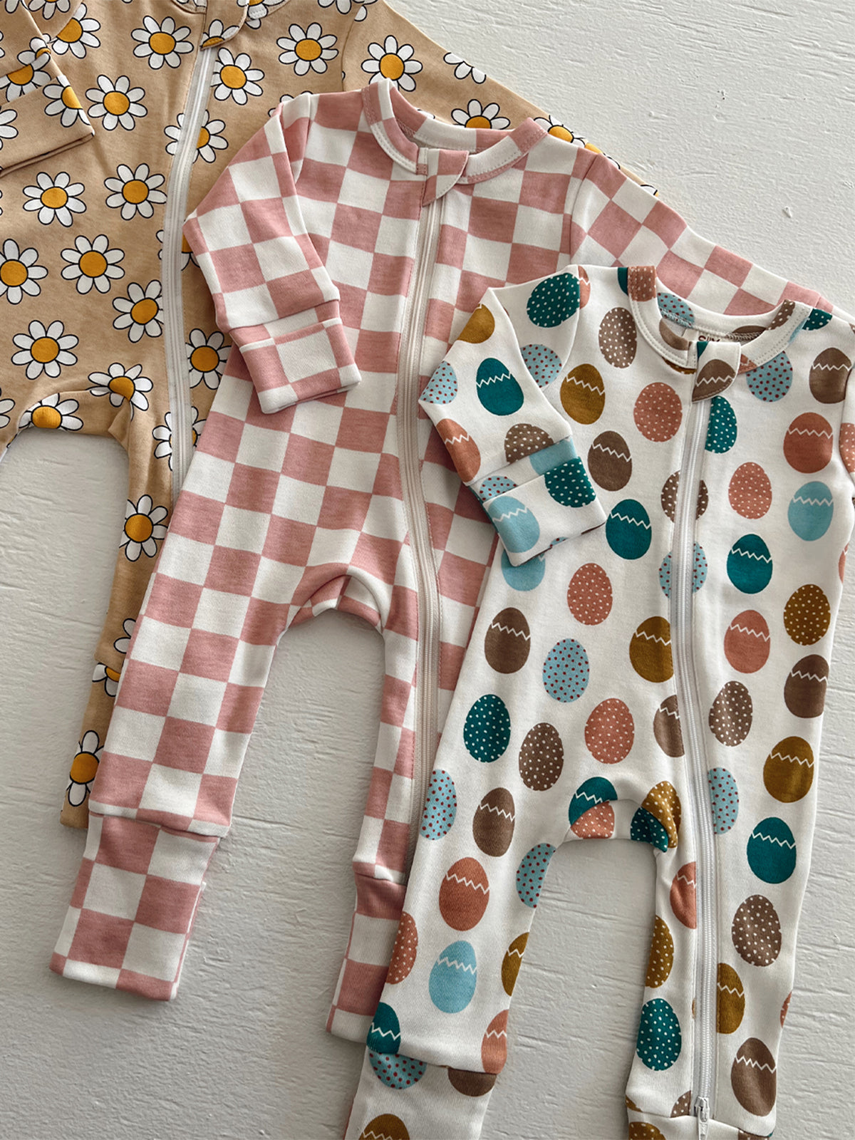 Three colorful baby onesies with floral, checkered, and egg patterns displayed on a white surface.