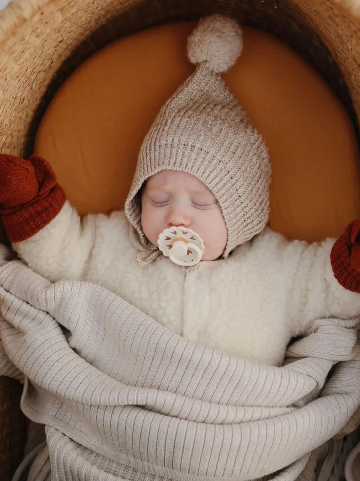 Sleeping baby in a cozy knitted hat and mittens, wrapped in a soft blanket in a woven basket.
