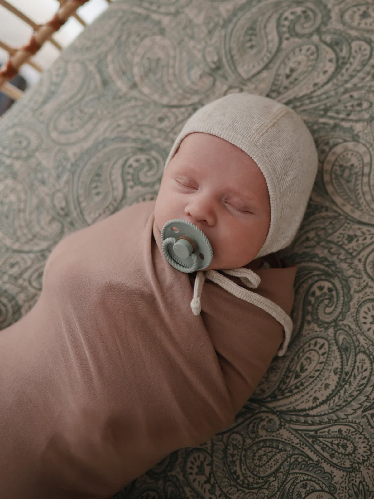 Sleeping baby swaddled in a brown blanket, wearing a light hat and pacifier, on a patterned green blanket.