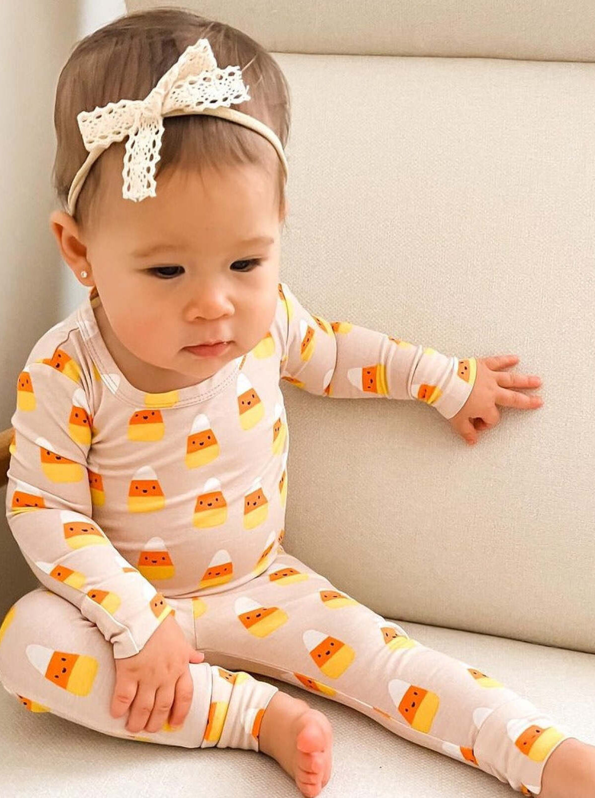 Toddler wearing candy corn-patterned outfit, seated on a light-colored sofa, with a lace headband.