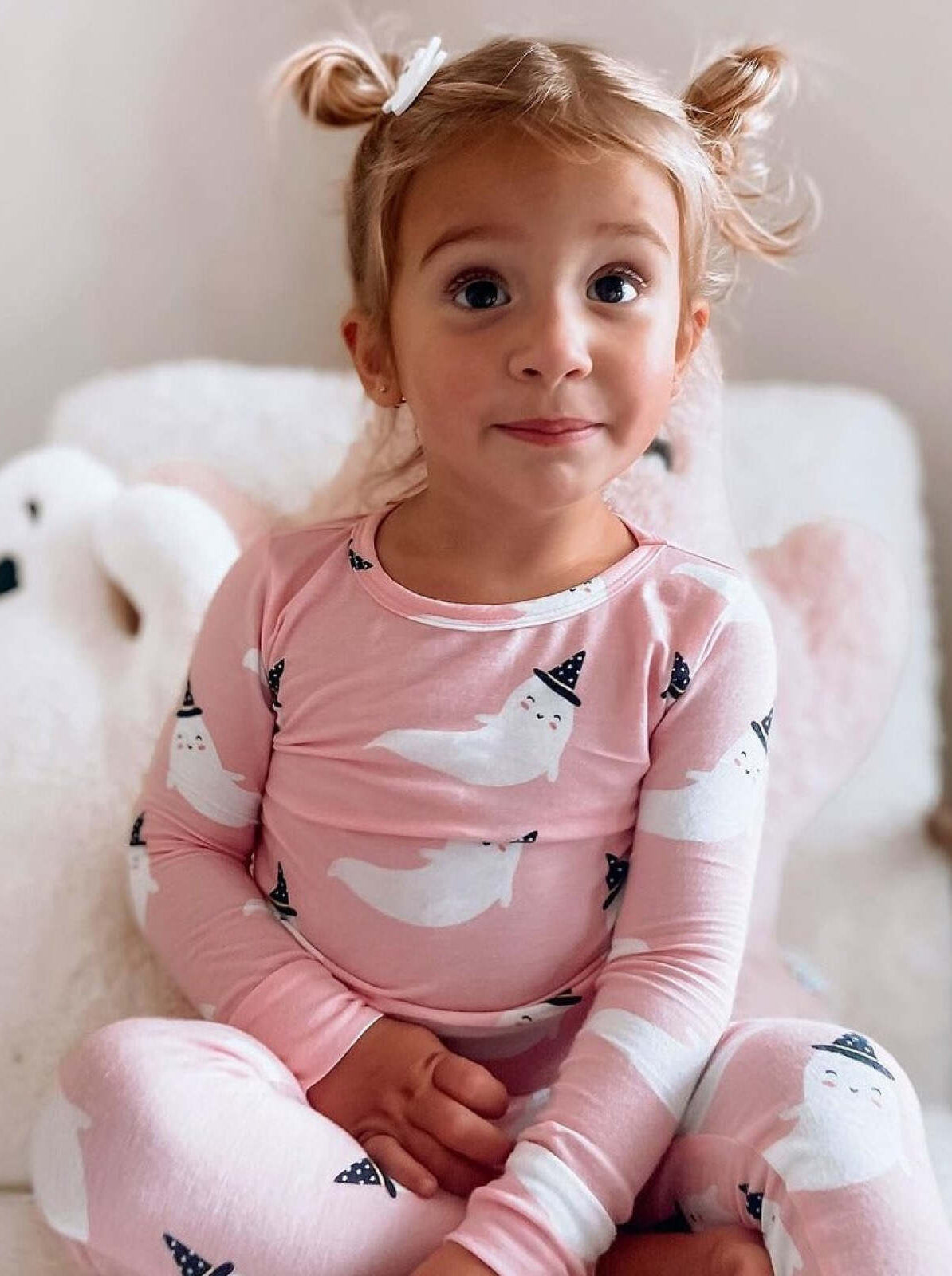 Young girl in pink ghost-patterned pajamas, smiling with playful hairstyle, sitting on soft pillows.