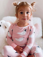 Young girl in pink ghost-patterned pajamas, smiling with playful hairstyle, sitting on soft pillows.