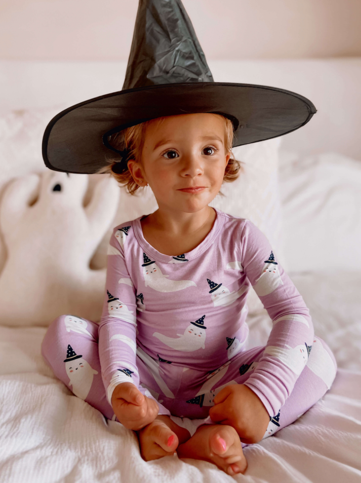 Smiling child in a witch hat and ghost pajamas, sitting on a bed with a plush ghost nearby.