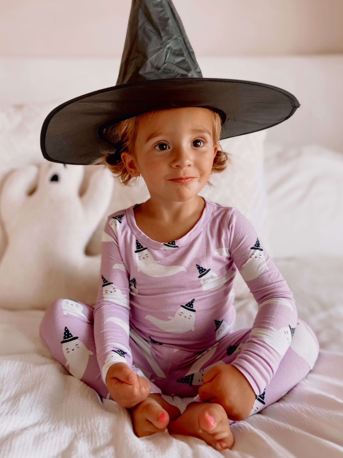 Smiling child in a witch hat and ghost pajamas, sitting on a bed with a plush ghost nearby.