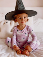 Smiling child in a witch hat and ghost pajamas, sitting on a bed with a plush ghost nearby.