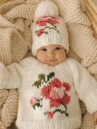 Smiling baby in a floral knit sweater and matching hat, resting on a cozy beige blanket.