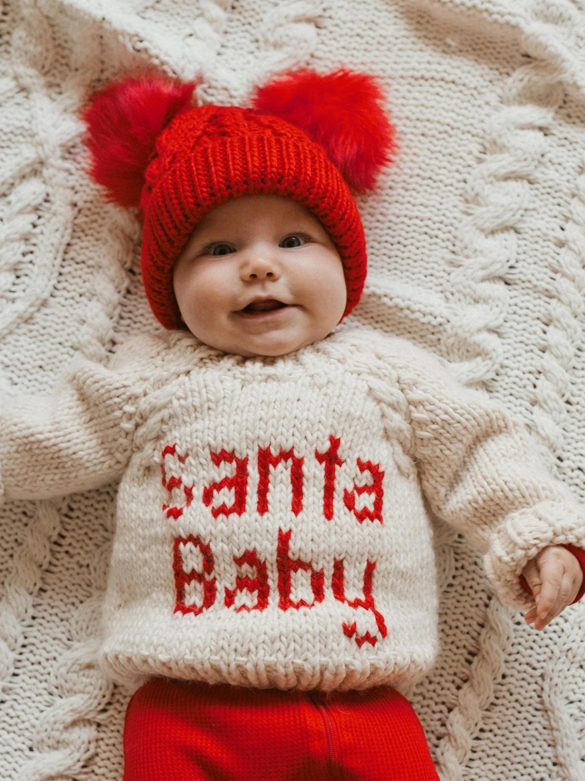 Smiling baby wearing a red pom-pom hat and a knitted sweater with "Santa Baby" on a cozy blanket.