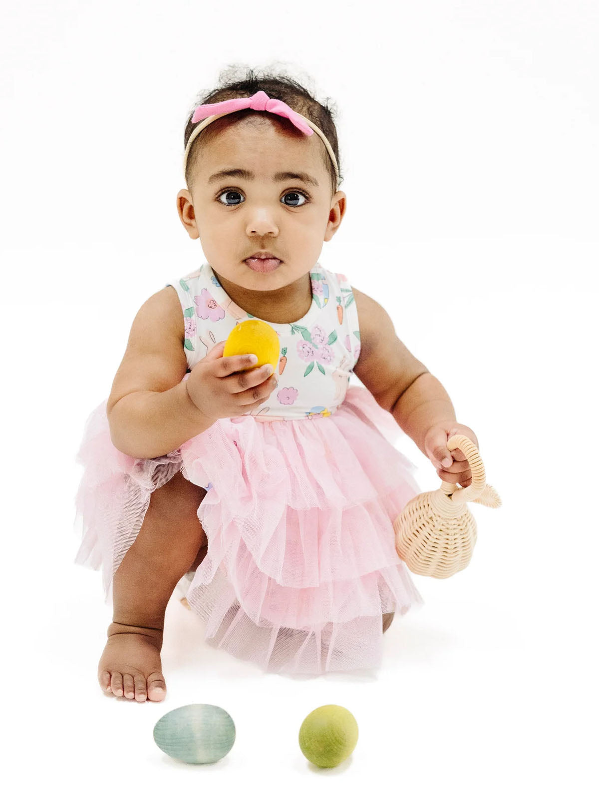 Toddler in a pink dress holding a yellow egg, with colorful wooden eggs nearby and a woven basket.