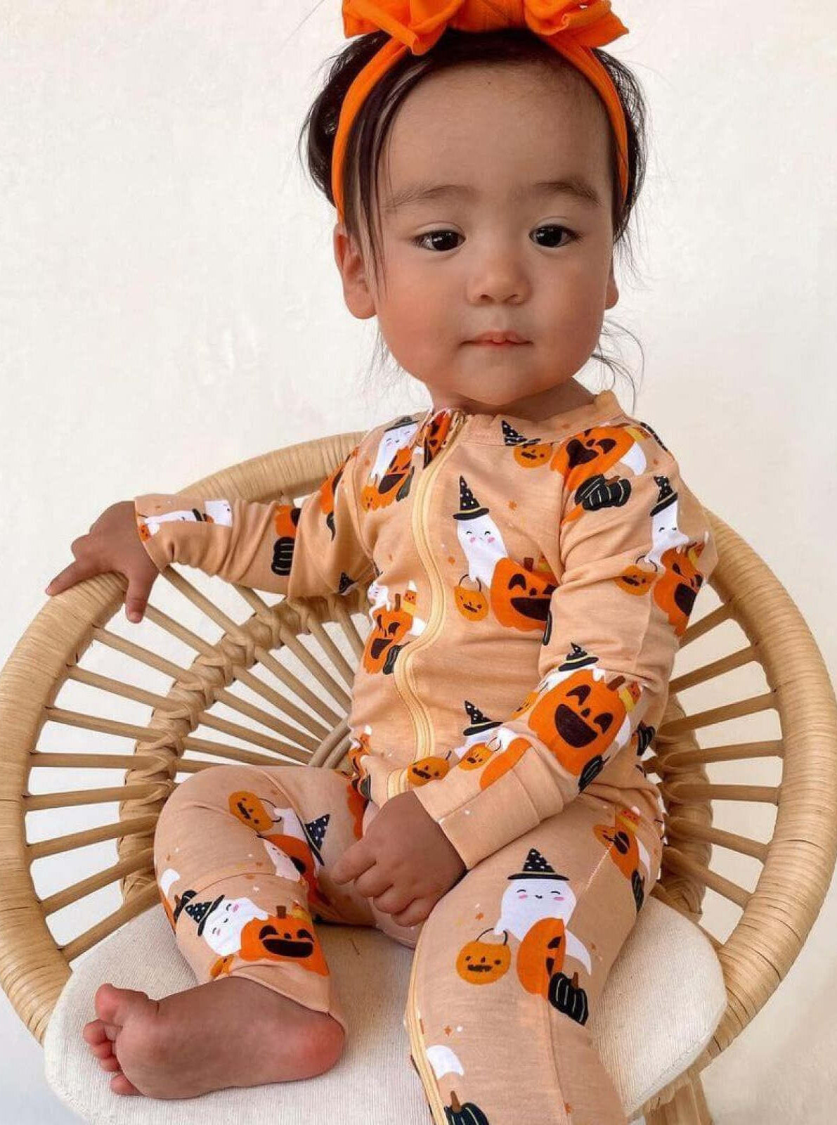 Toddler in an orange Halloween-themed outfit poses on a stylish chair with a big orange bow in her hair.