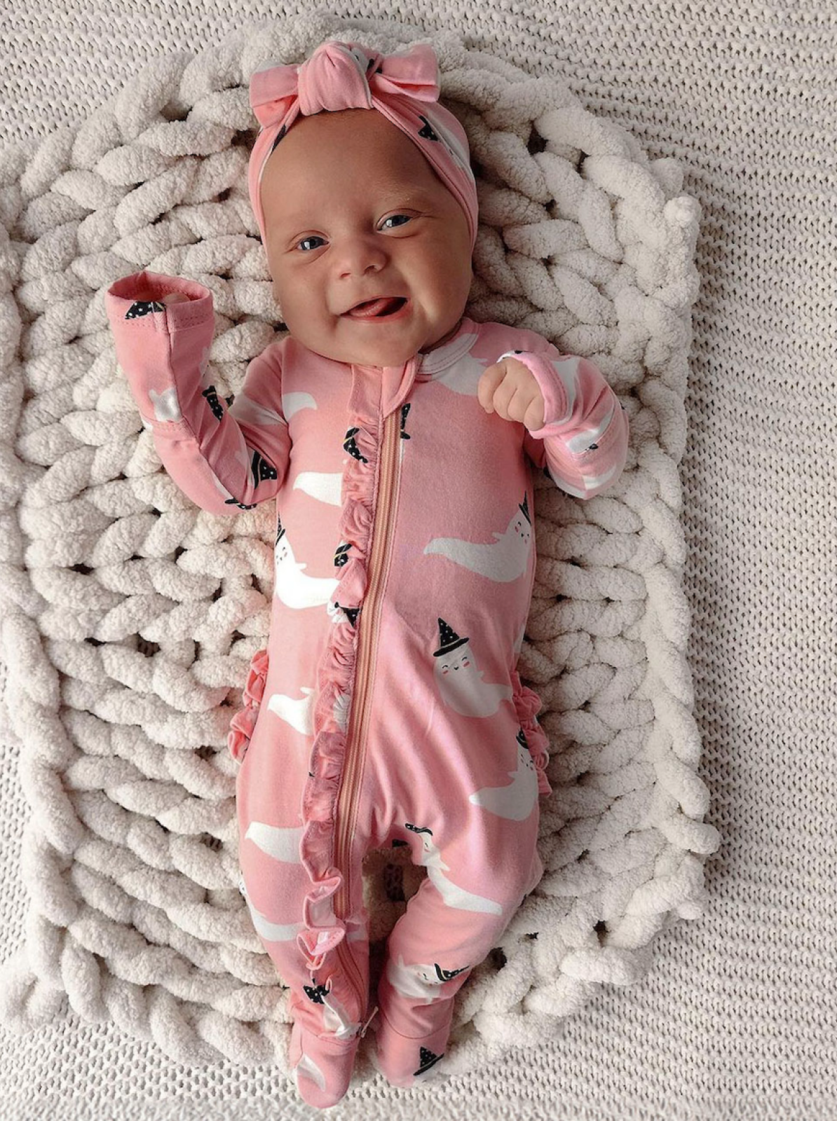 Smiling baby in a pink ghost-patterned onesie and headband, lying on a textured blanket.