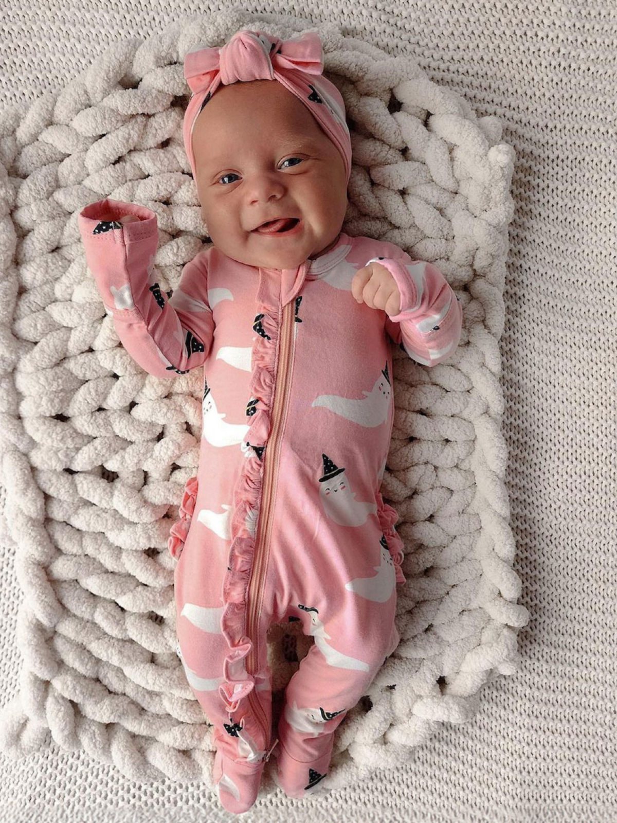 Smiling baby in a pink ghost-patterned onesie and headband, lying on a textured blanket.