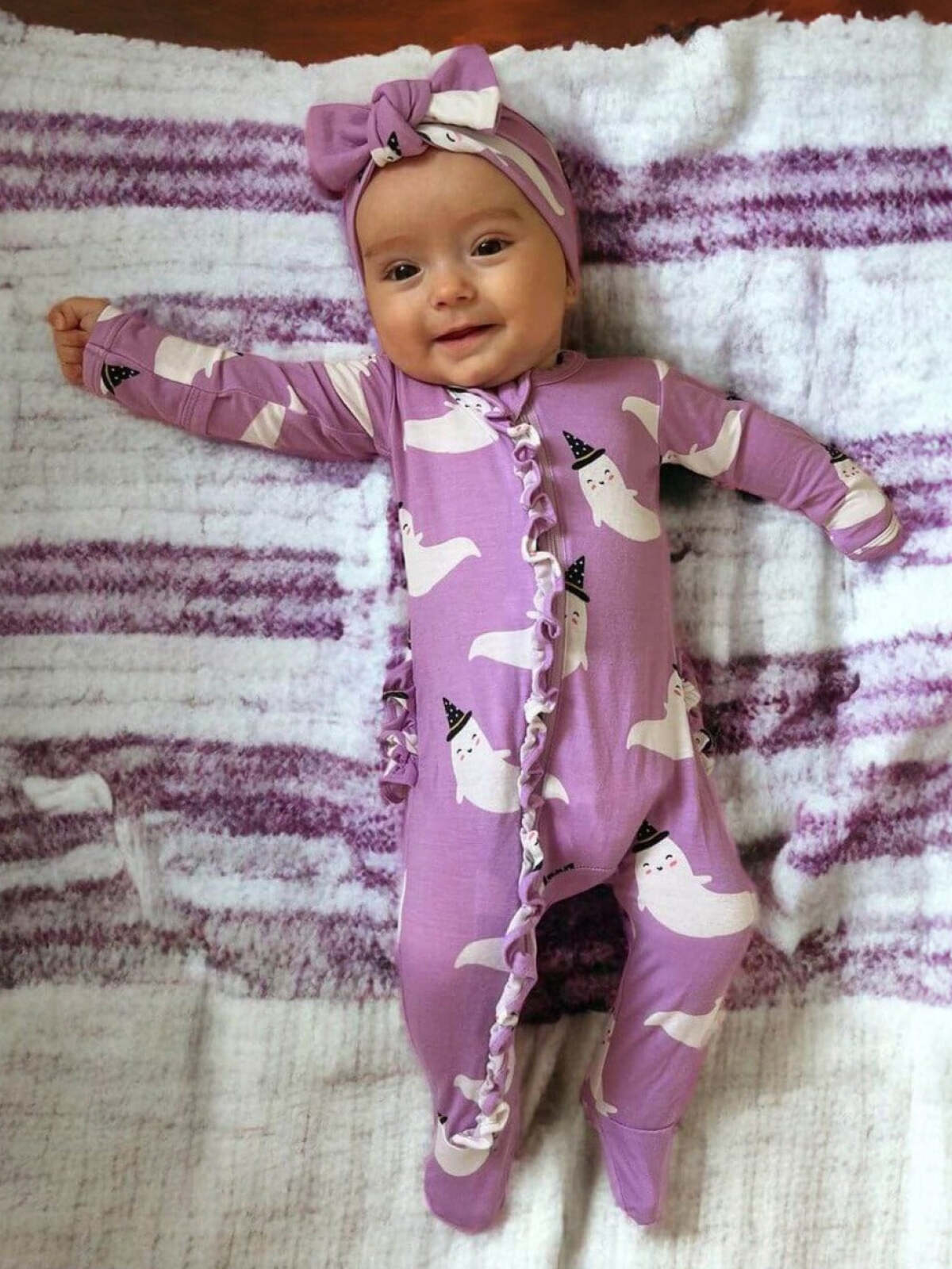 Smiling baby in a purple ghost-patterned onesie, wearing a matching headband, lying on a soft blanket.