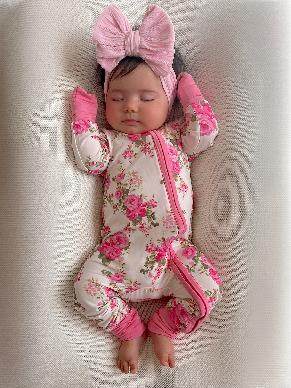Sleeping baby in floral onesie with pink accents and a large bow headband on a soft, light background.