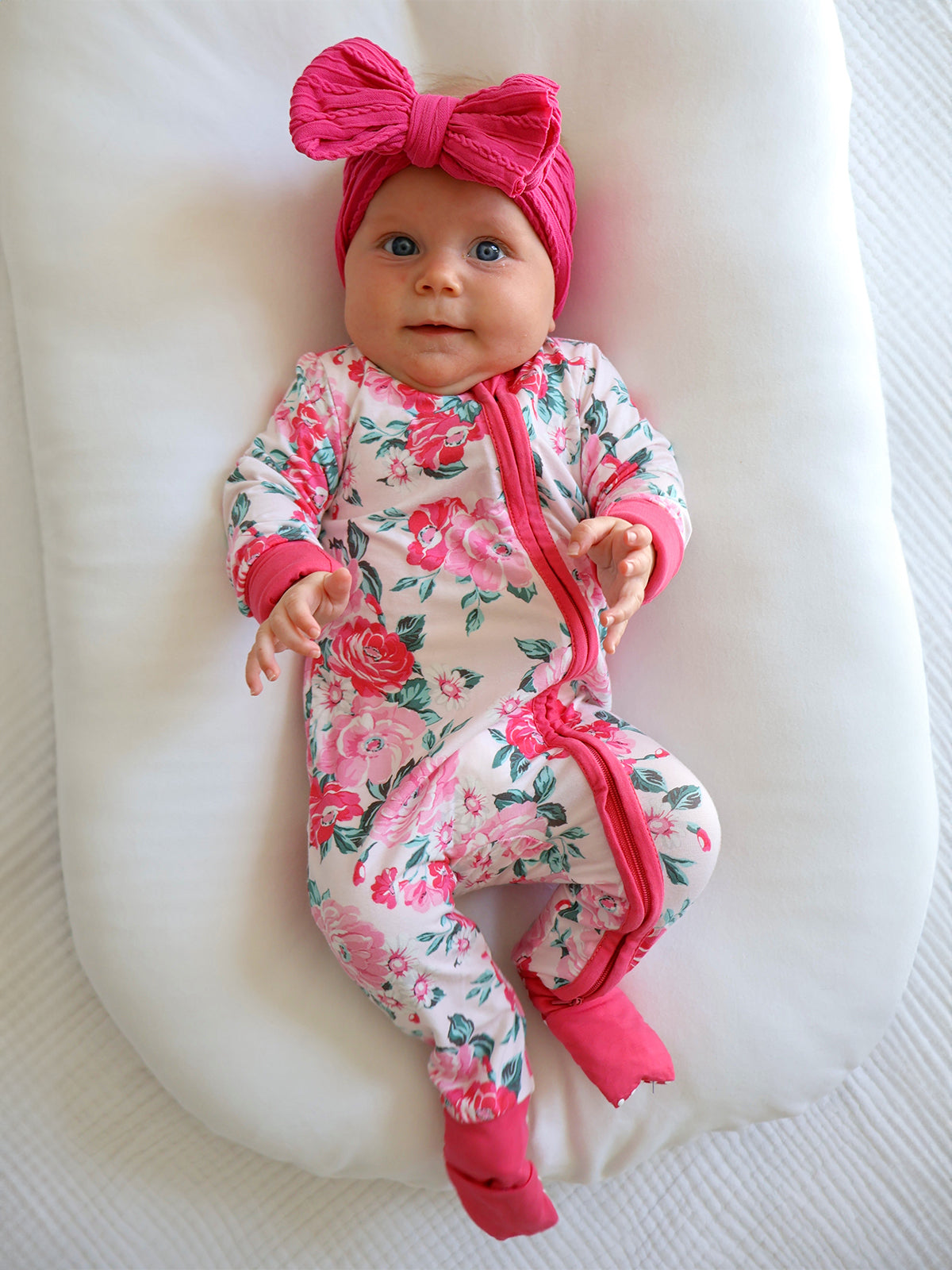 Smiling baby in floral pajamas and a pink bow, lying on a soft white surface.