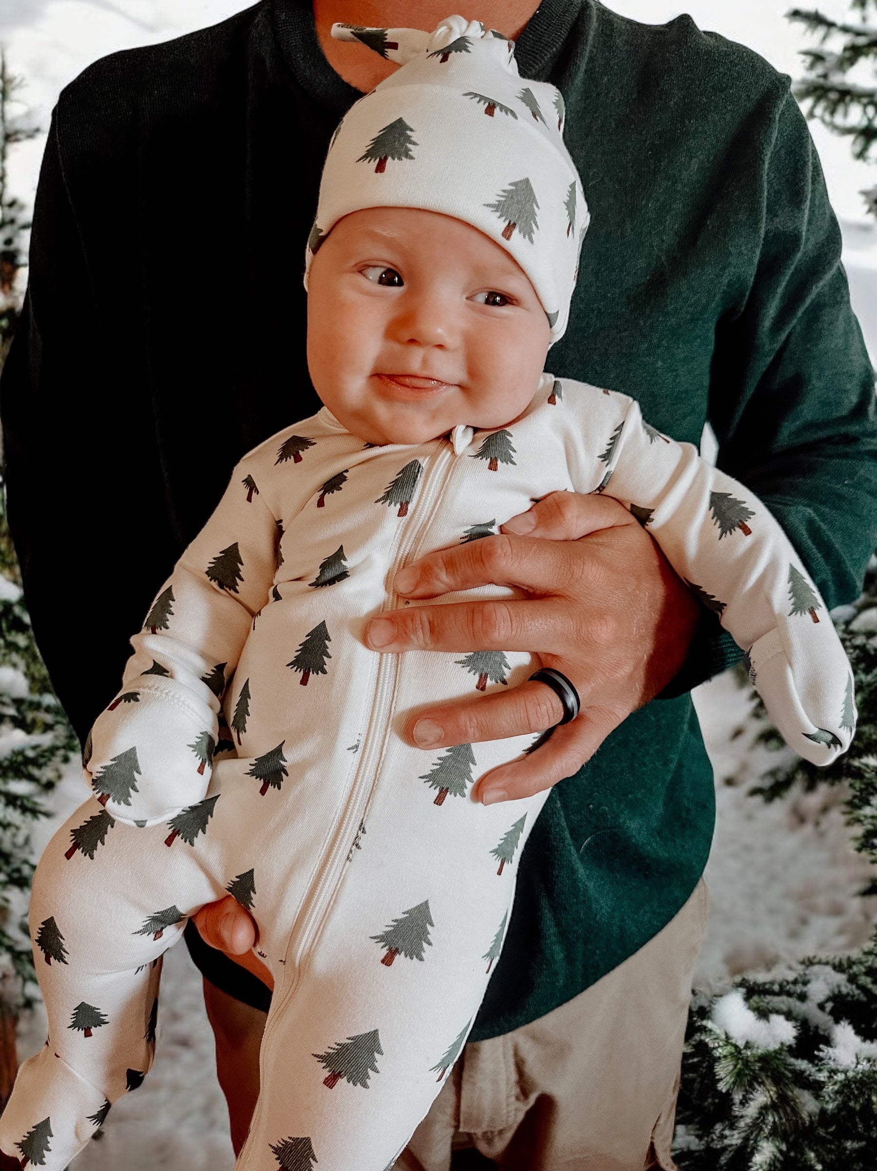 Infant wearing tree-patterned pajamas, smiling while being held by an adult in a snowy forest setting.