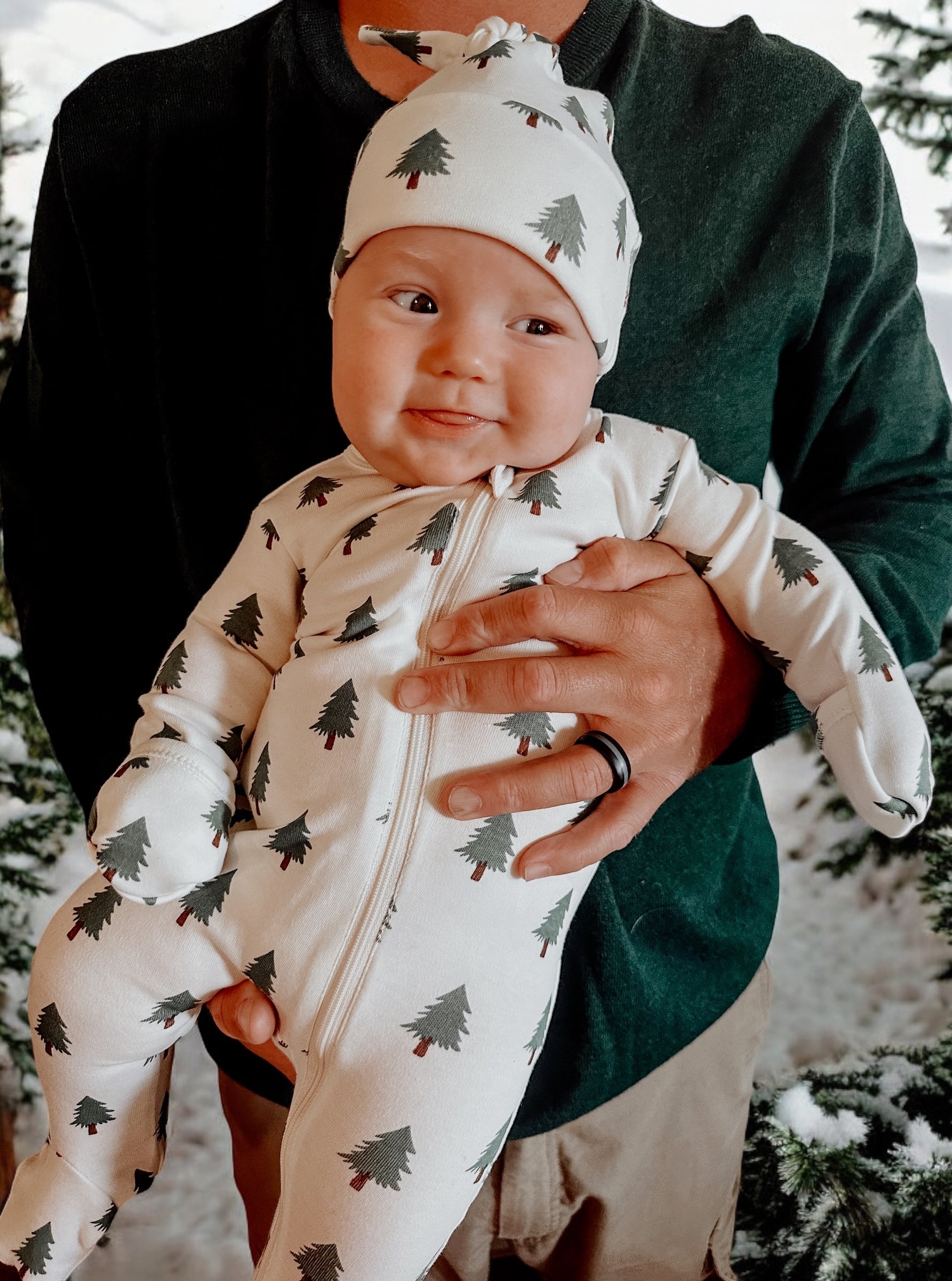 Infant wearing tree-patterned pajamas, smiling while being held by an adult in a snowy forest setting.