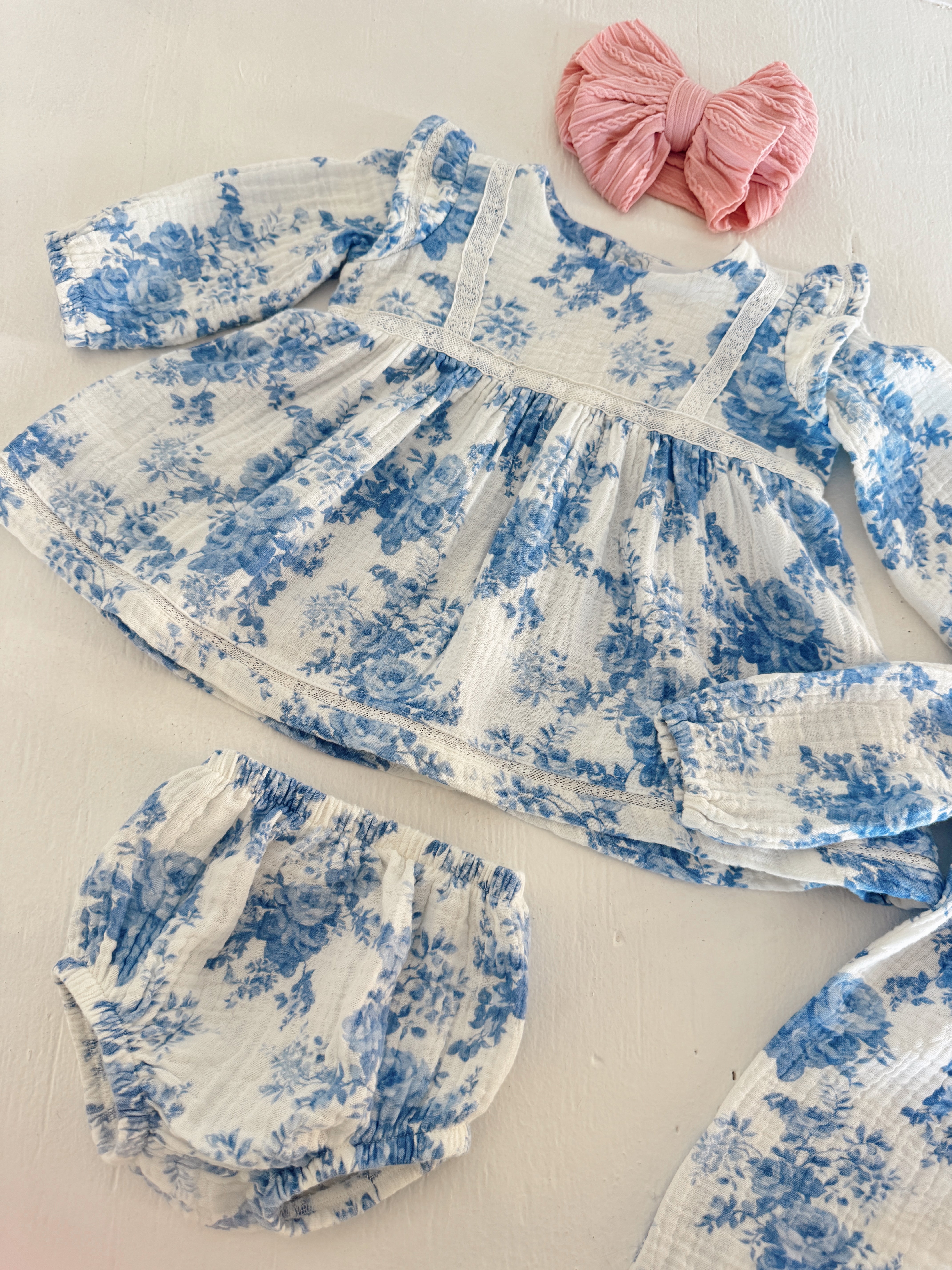 Baby dress and bloomers in white with blue floral print, paired with a pink bow on a light surface.
