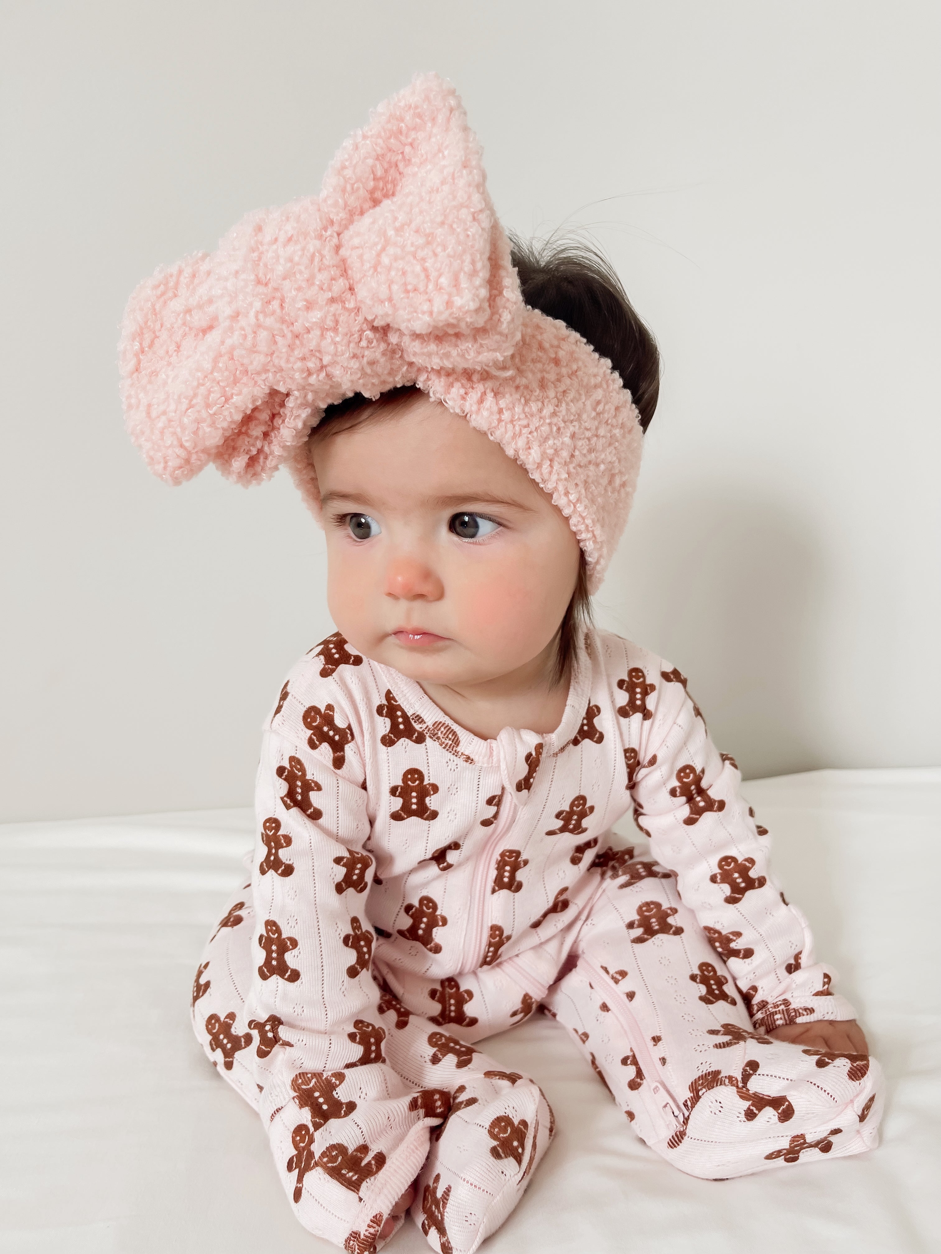 Infant girl with a pink bow headband and gingerbread-patterned pajamas sitting on a white surface, looking off to the side.