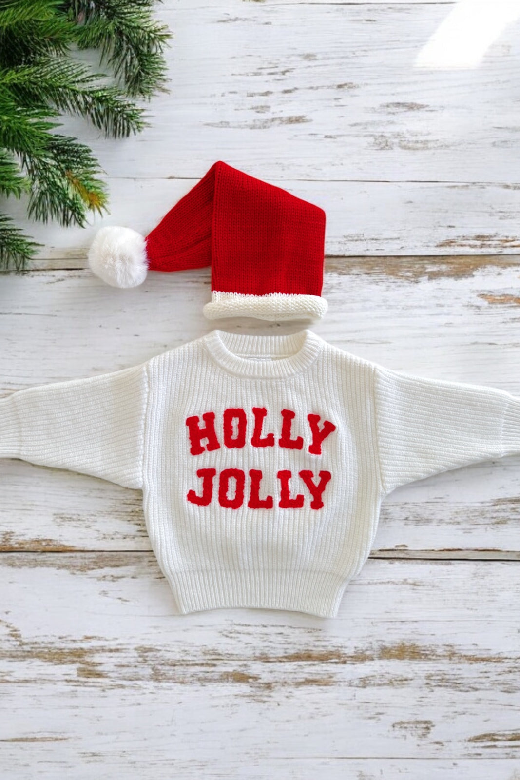 White sweater with "HOLLY JOLLY" in red, alongside a red Christmas hat, placed on a wooden background.