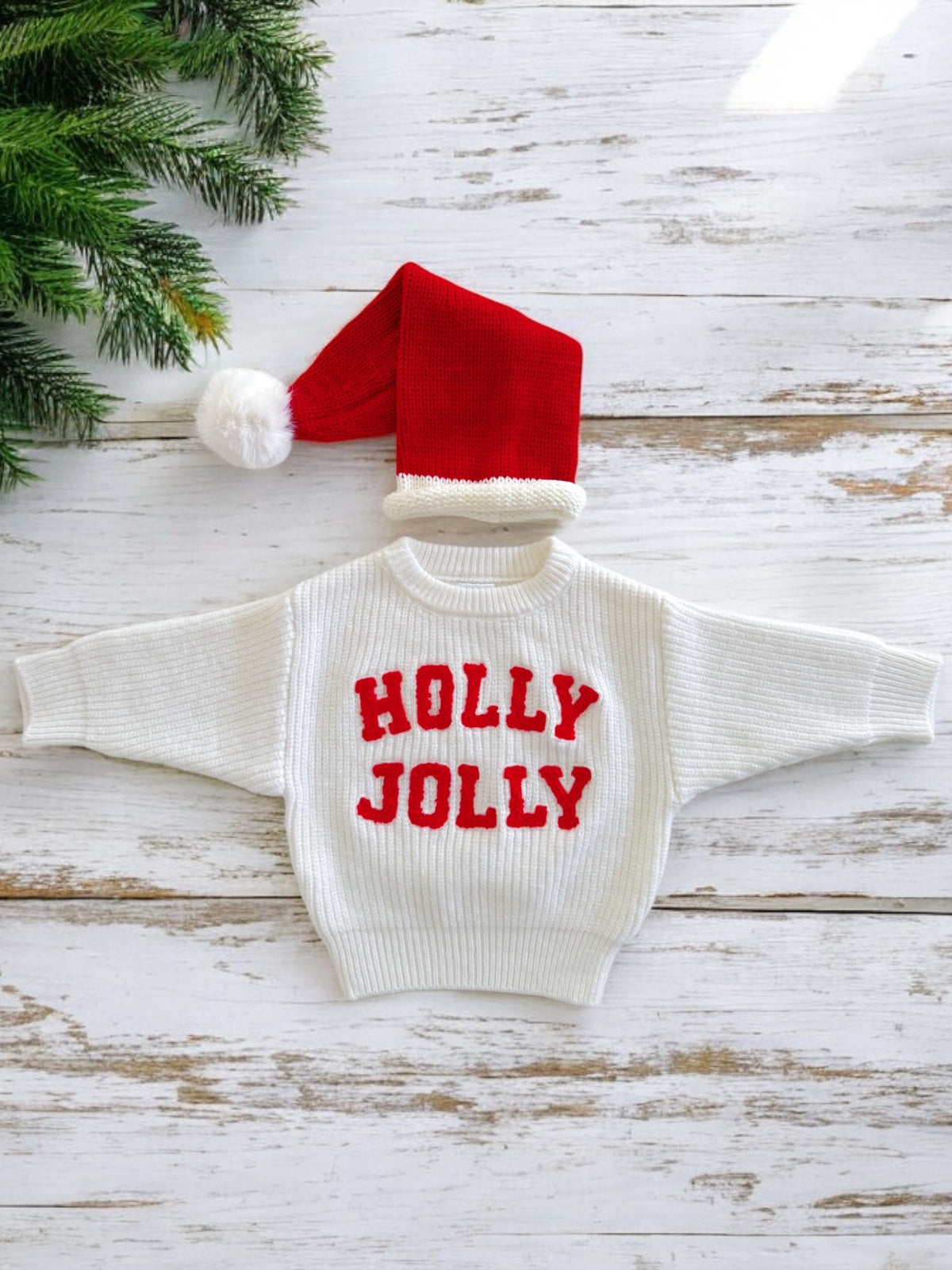 White sweater with "HOLLY JOLLY" in red, alongside a red Christmas hat, placed on a wooden background.