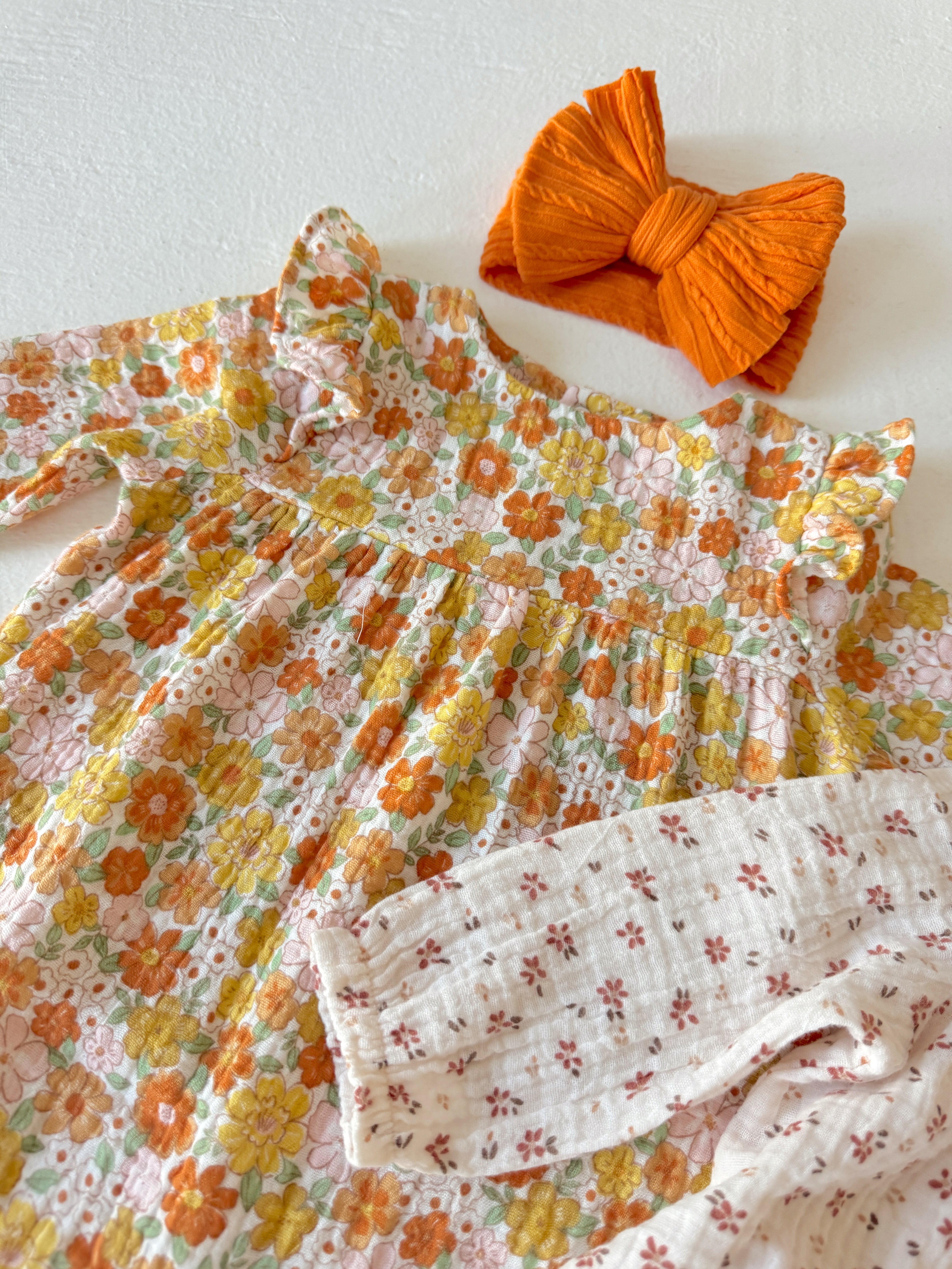 Floral baby dress with ruffled collar and a matching orange bow on a white background.