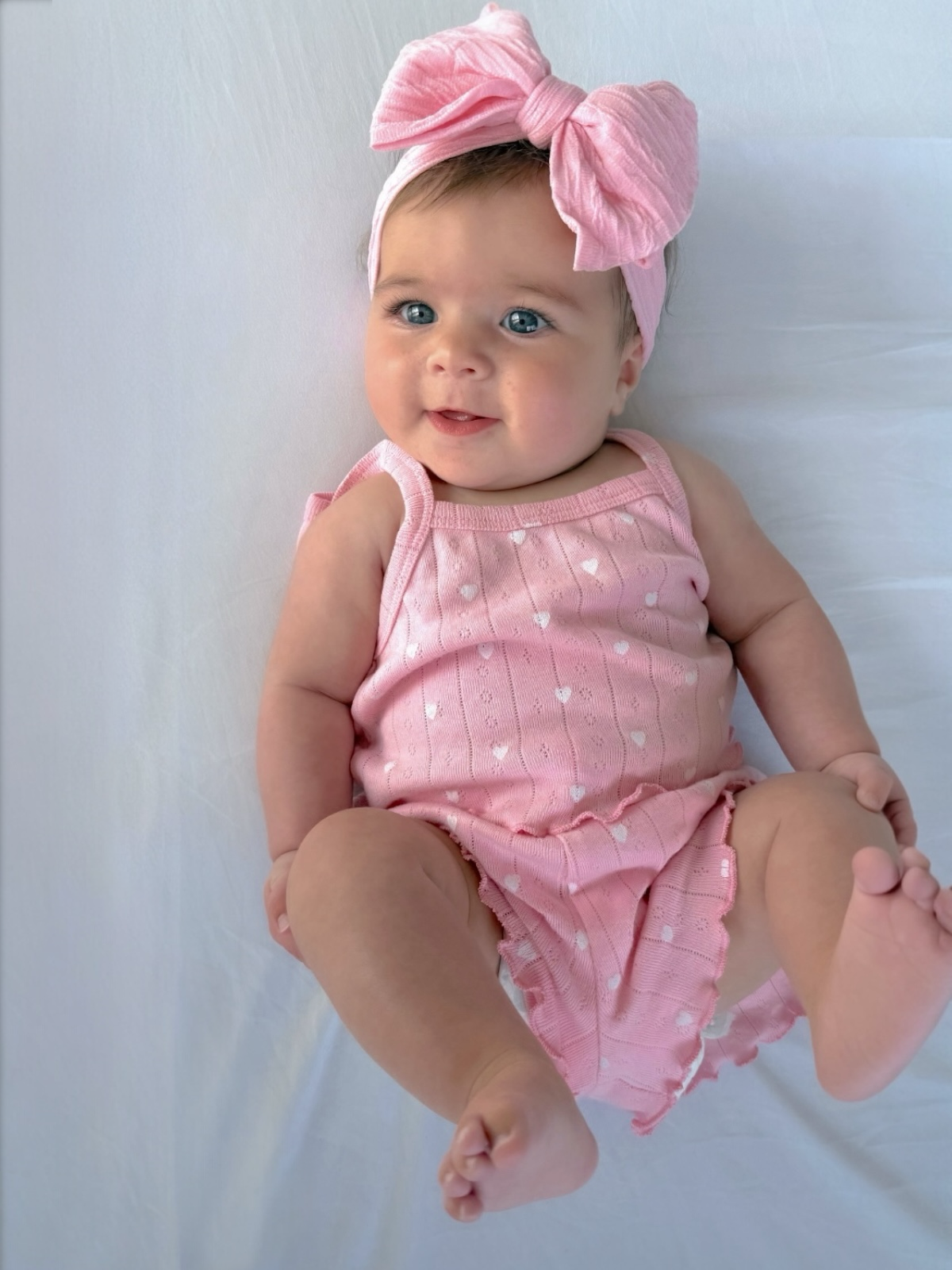 Smiling baby wearing a pink outfit and headband, lying on a white surface with legs outstretched.