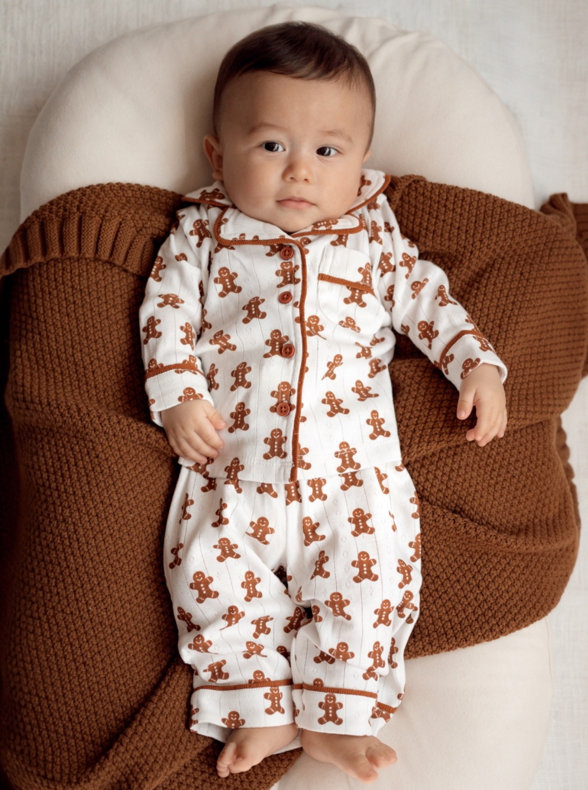 Baby in gingerbread-themed pajamas resting on a cozy brown blanket, with a calm expression.