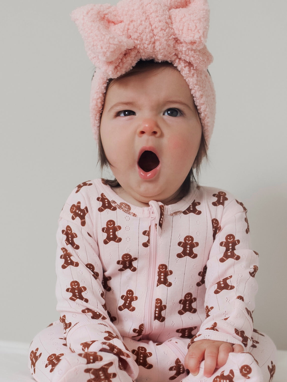 Baby in gingerbread-patterned pajamas yawns while wearing a fluffy pink headband.