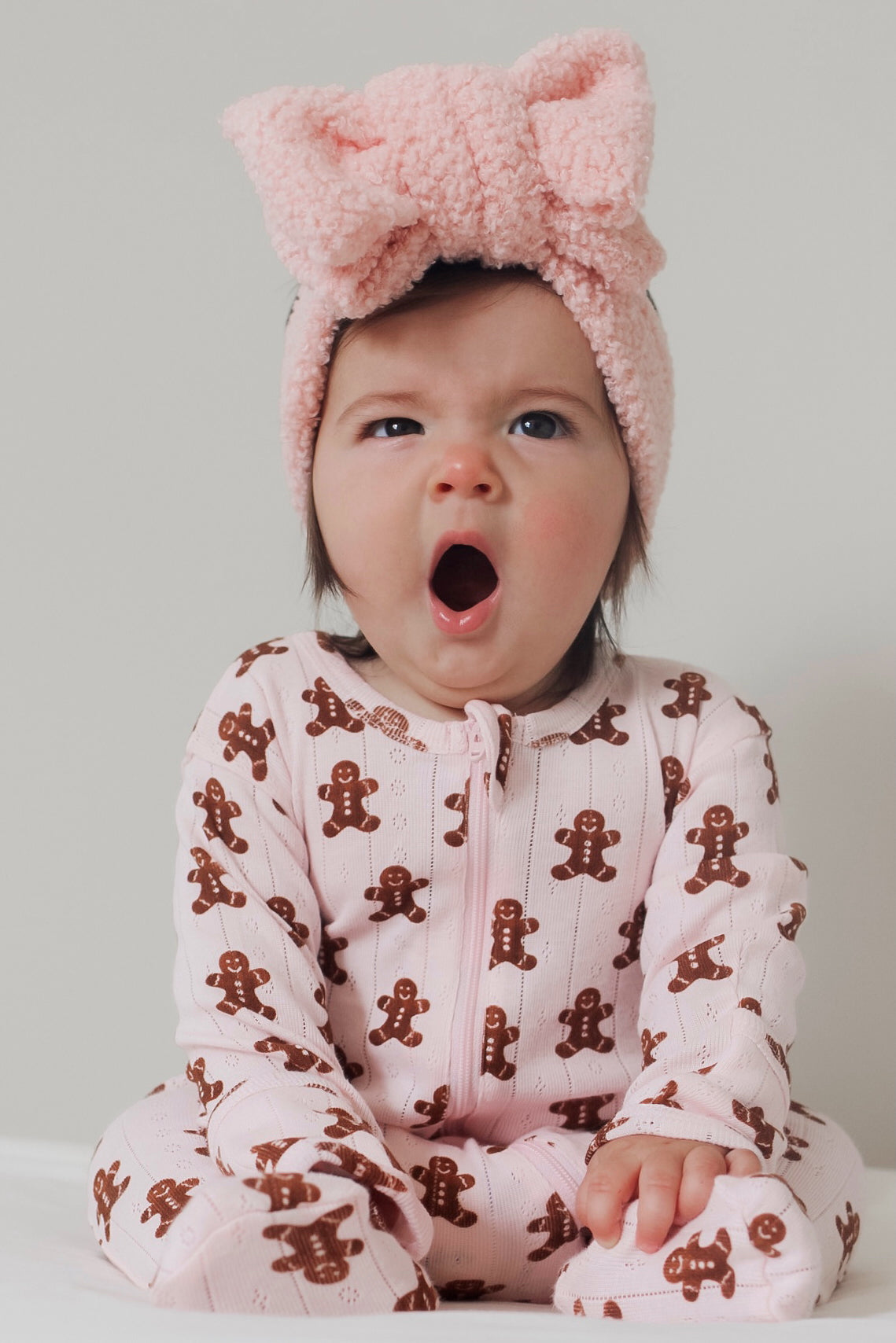Baby in gingerbread-patterned pajamas yawns while wearing a fluffy pink headband.